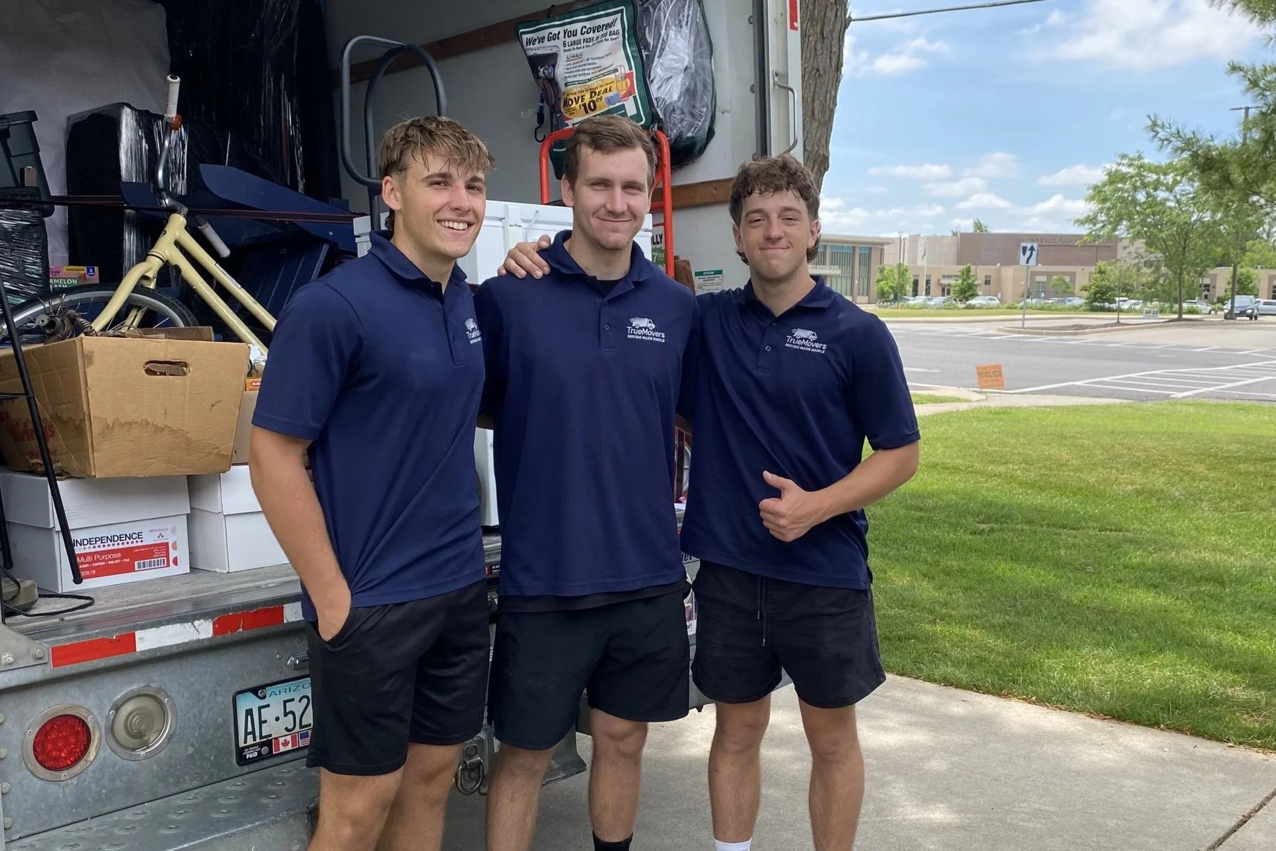 Three young men wearing matching navy blue polo shirts and black shorts standing in front of a moving truck. They are smiling and posing for the photo with one man having his arm around another. The moving truck is filled with boxes, a bicycle, and various other items. The background shows a parking lot, some trees, and a building under a partly cloudy sky.