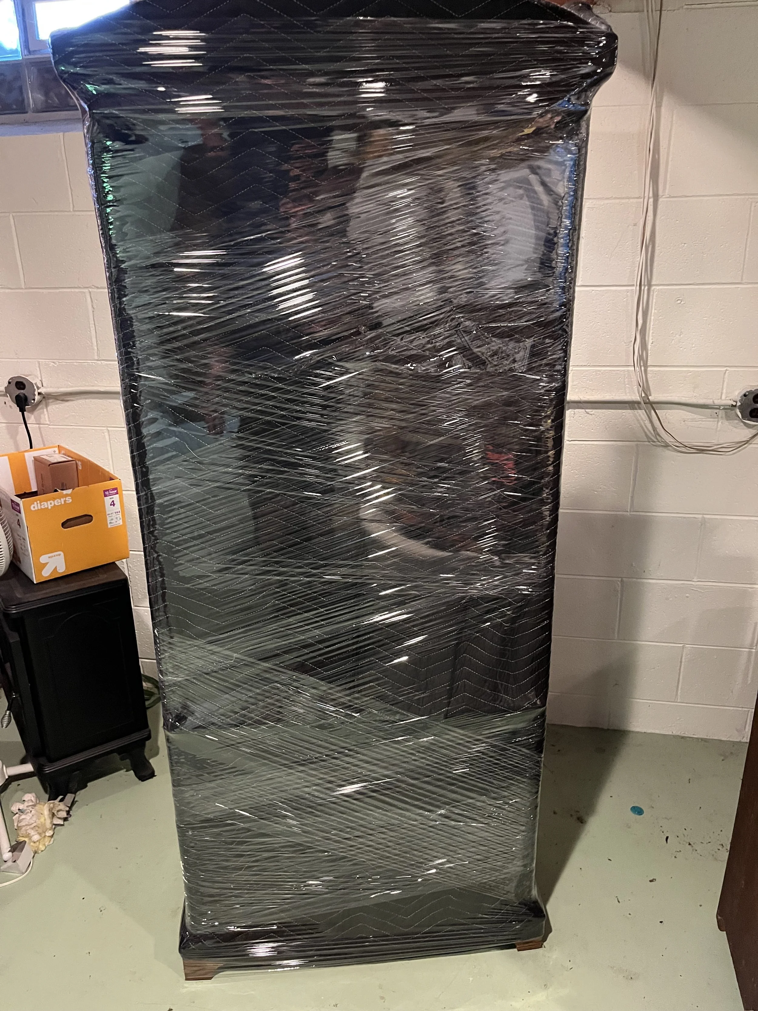 A black refrigerator wrapped in plastic wrap standing in a room with fluorescent lighting and a white brick wall.