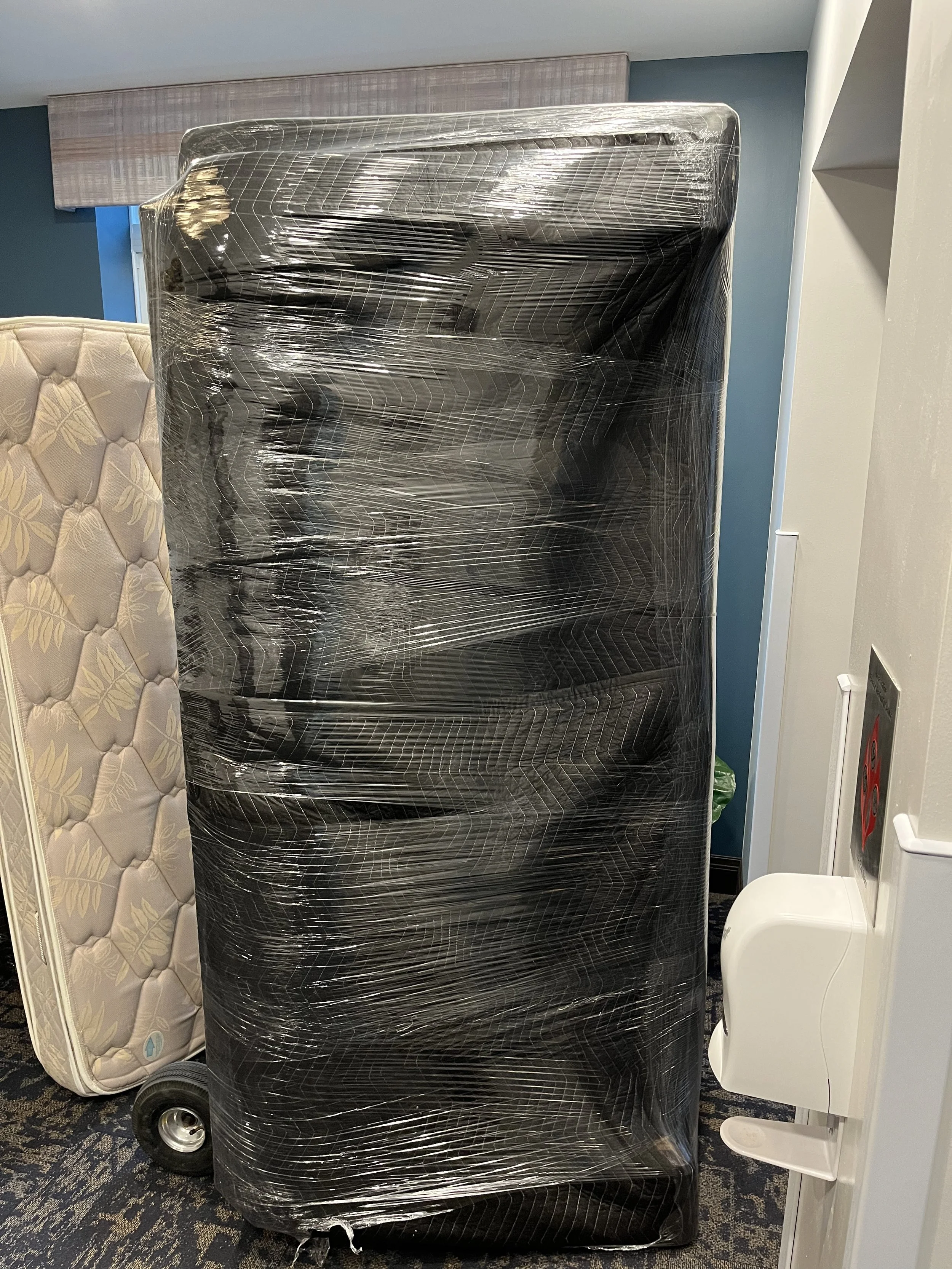 A large piece of furniture wrapped in plastic sitting in a room, with a mattress to the left and a white chair on the right. The room has blue and white walls, and a patterned carpet.