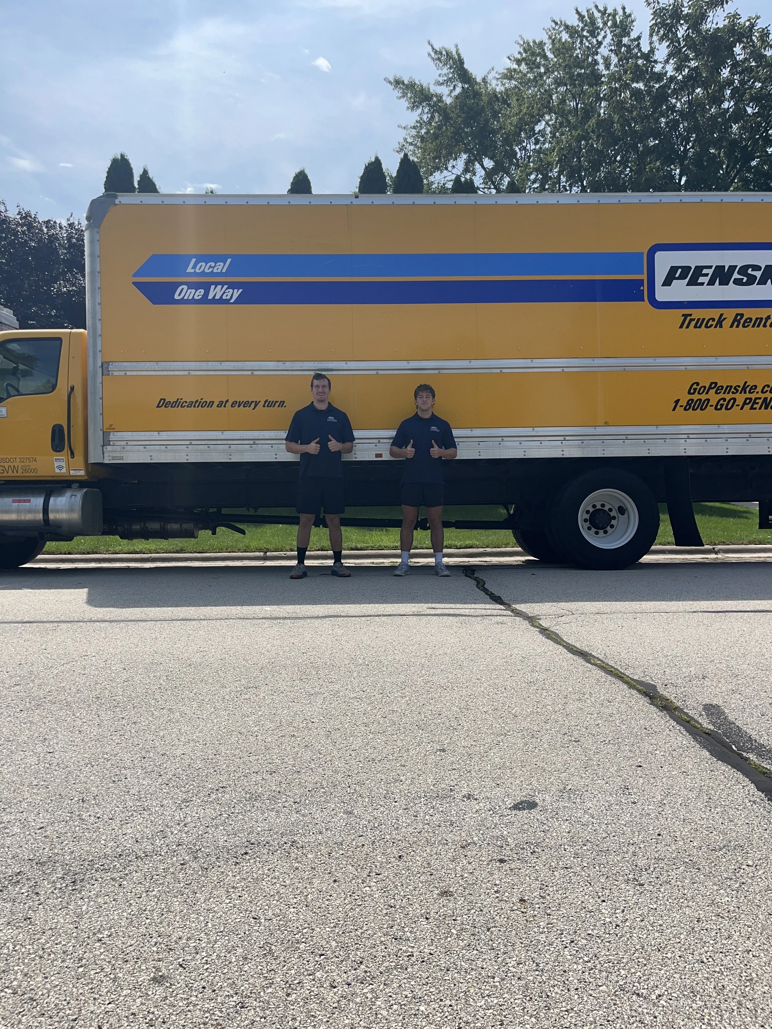 A yellow Penske truck with two individuals standing in front of it, giving thumbs-up signs outdoors on a sunny day. There are trees in the background and an asphalt road beneath.
