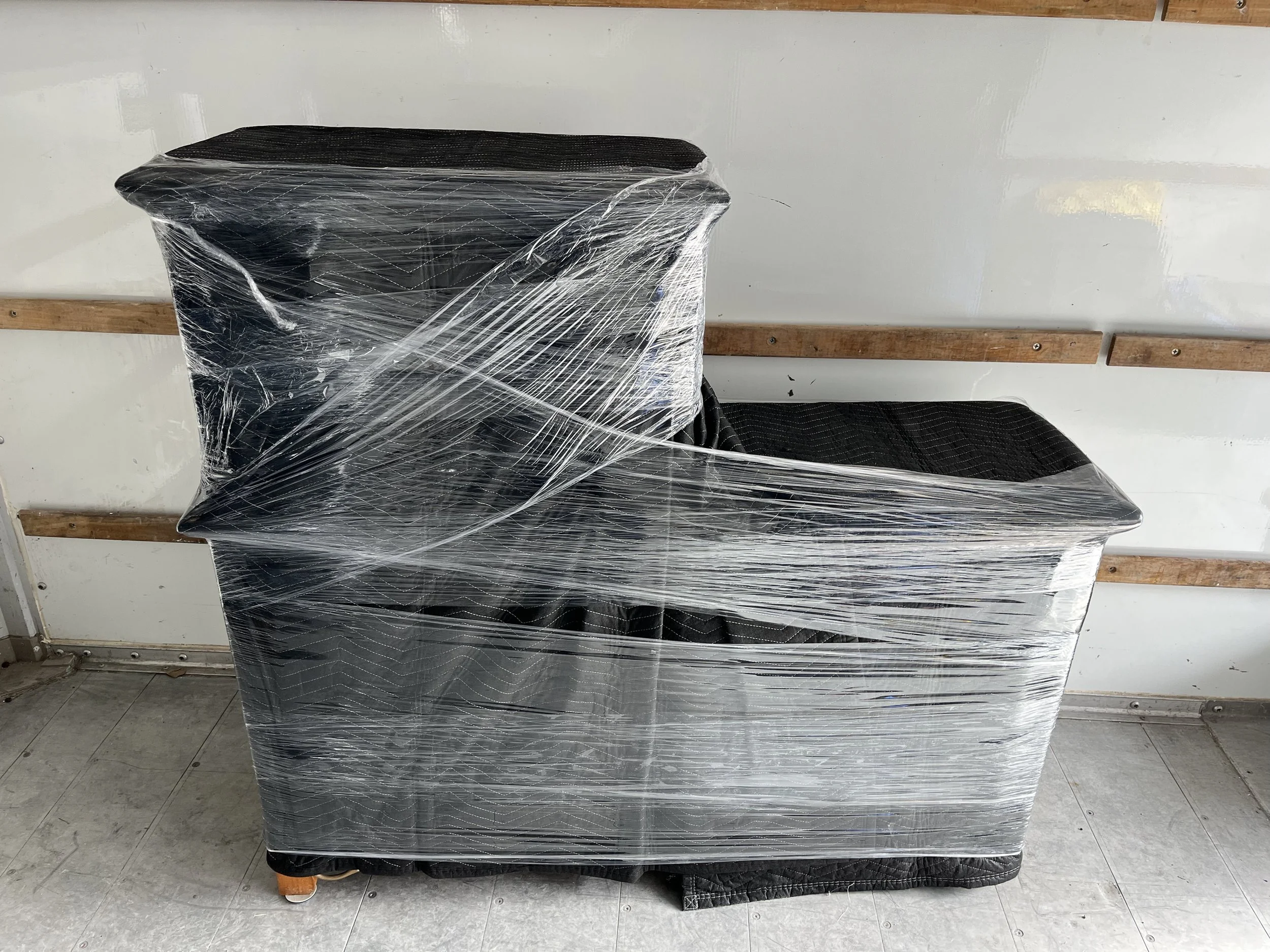 Wrapped black furniture or appliance covered in clear plastic stretch wrap, placed against a wall with wooden strips and a tiled floor.