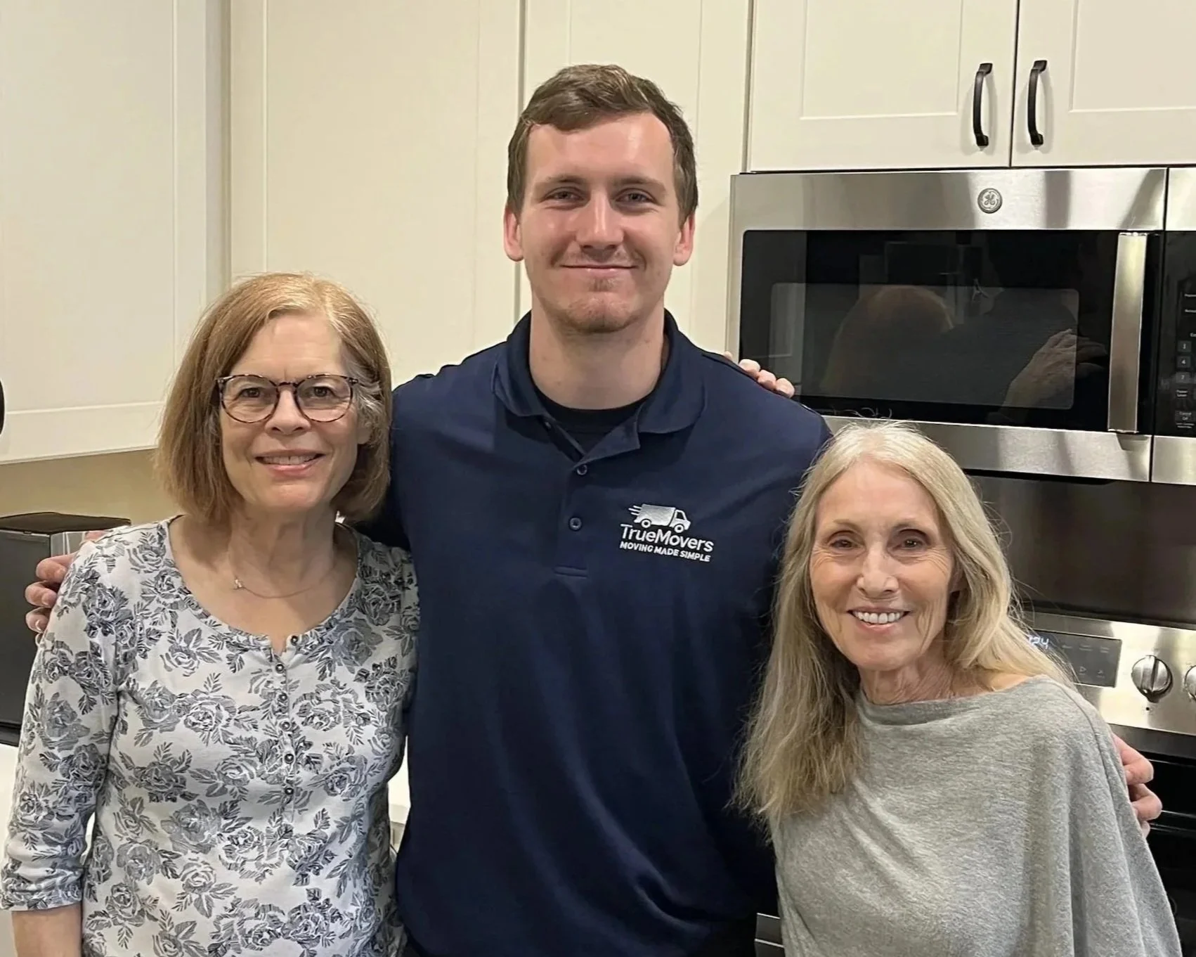 Three women and one man smiling for a photo in a kitchen, with white cabinets and a stainless steel microwave behind them. The man is wearing a navy blue shirt with a company logo.