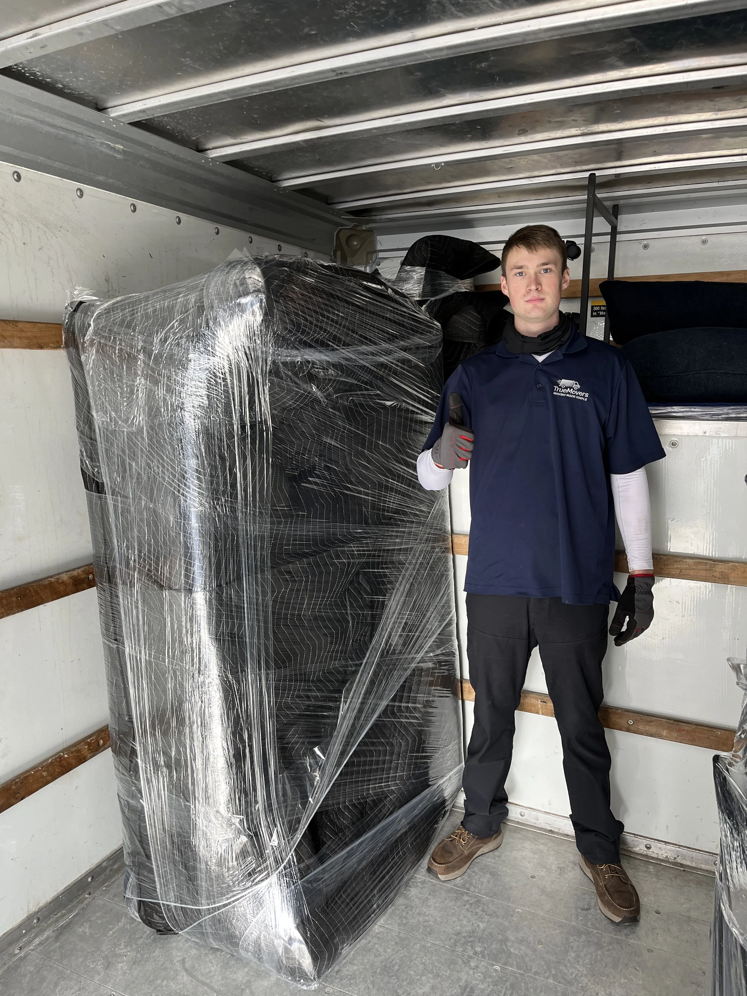 A man standing inside a moving truck next to a large item wrapped in plastic, giving a thumbs up.