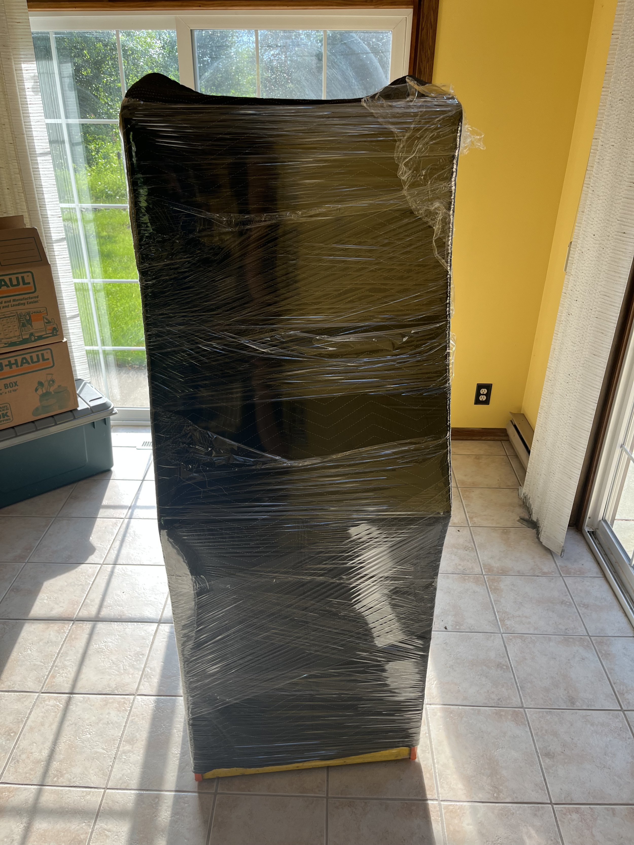 A large black box wrapped in plastic wrap standing upright on a tiled floor near a sliding glass door, with cardboard boxes to the side and a window showing greenery outside.