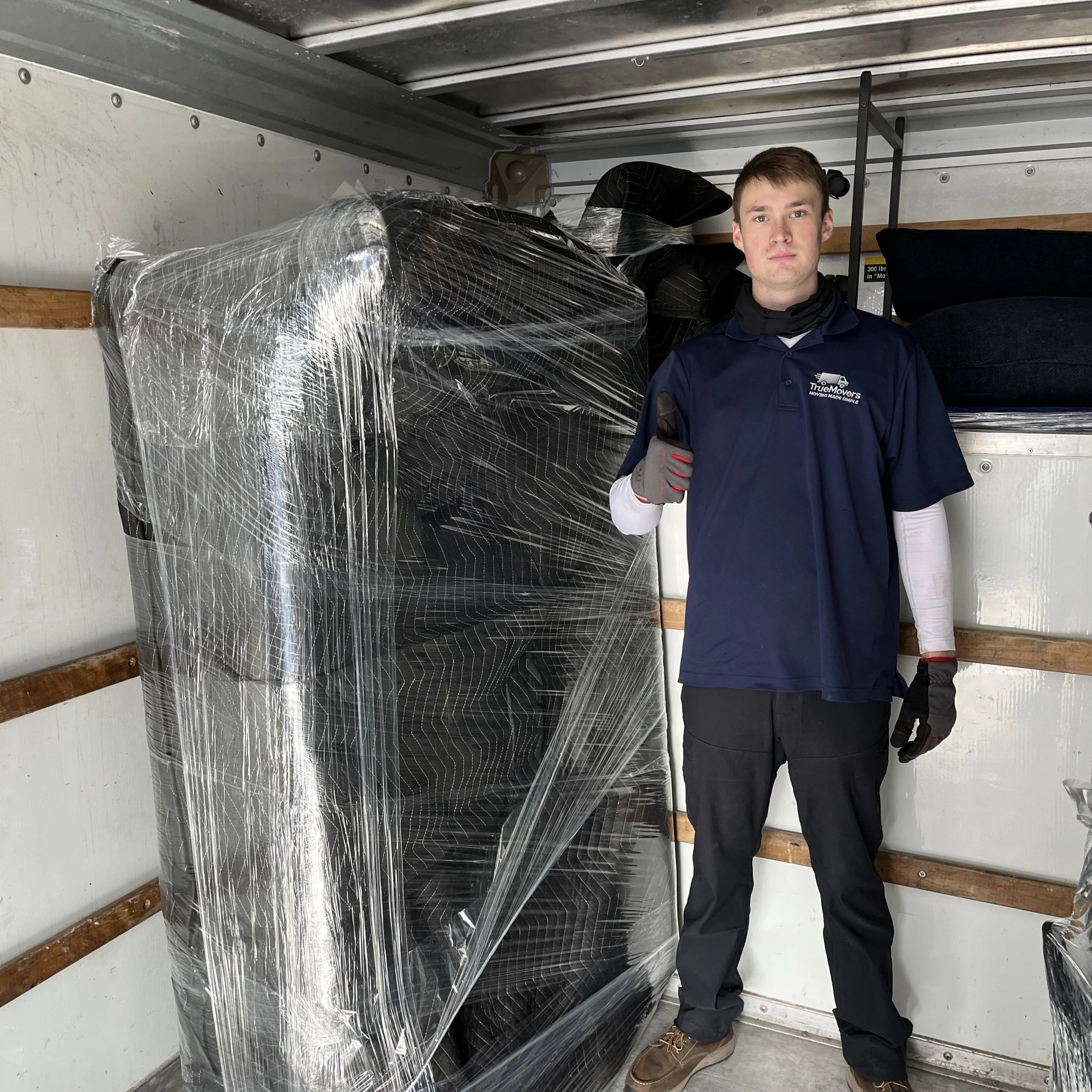 A young man in a blue uniform standing inside a truck, giving a thumbs up, with packed furniture wrapped in plastic behind him.