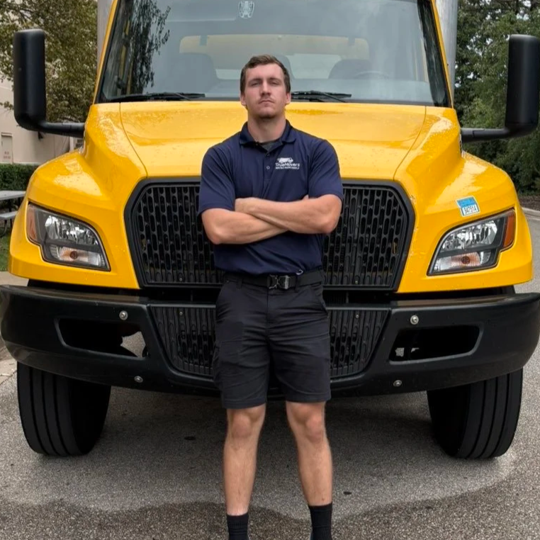 A man standing with crossed arms in front of a large yellow commercial truck, wearing a navy polo shirt and black shorts.