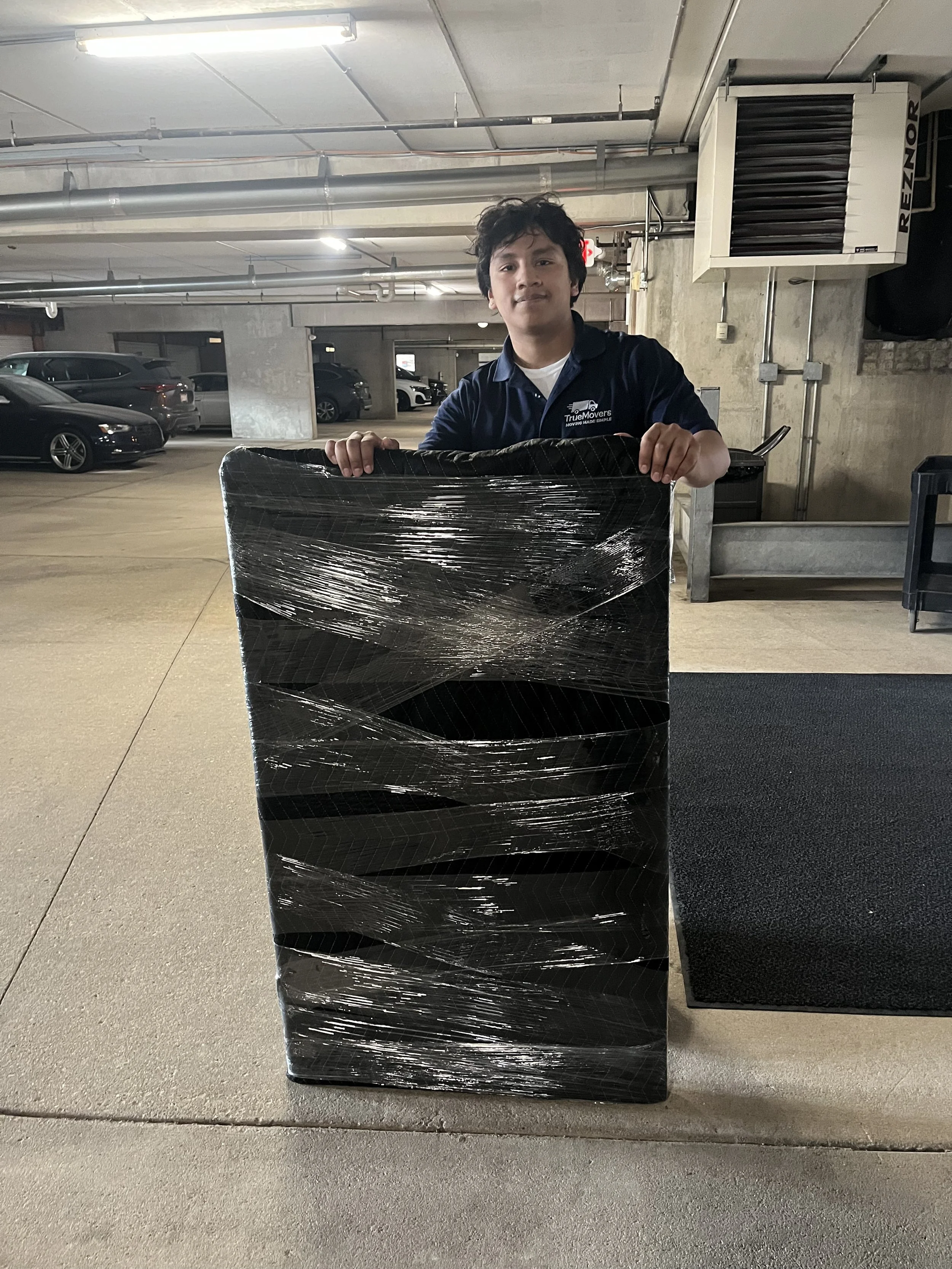 A young man in a dark blue shirt with a company logo is holding a large wrapped pallet in a parking garage.