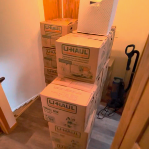 Stack of four U-Haul boxes in a hallway, with a white paper bag on top, and cleaning supplies including a mop and vacuum nearby.