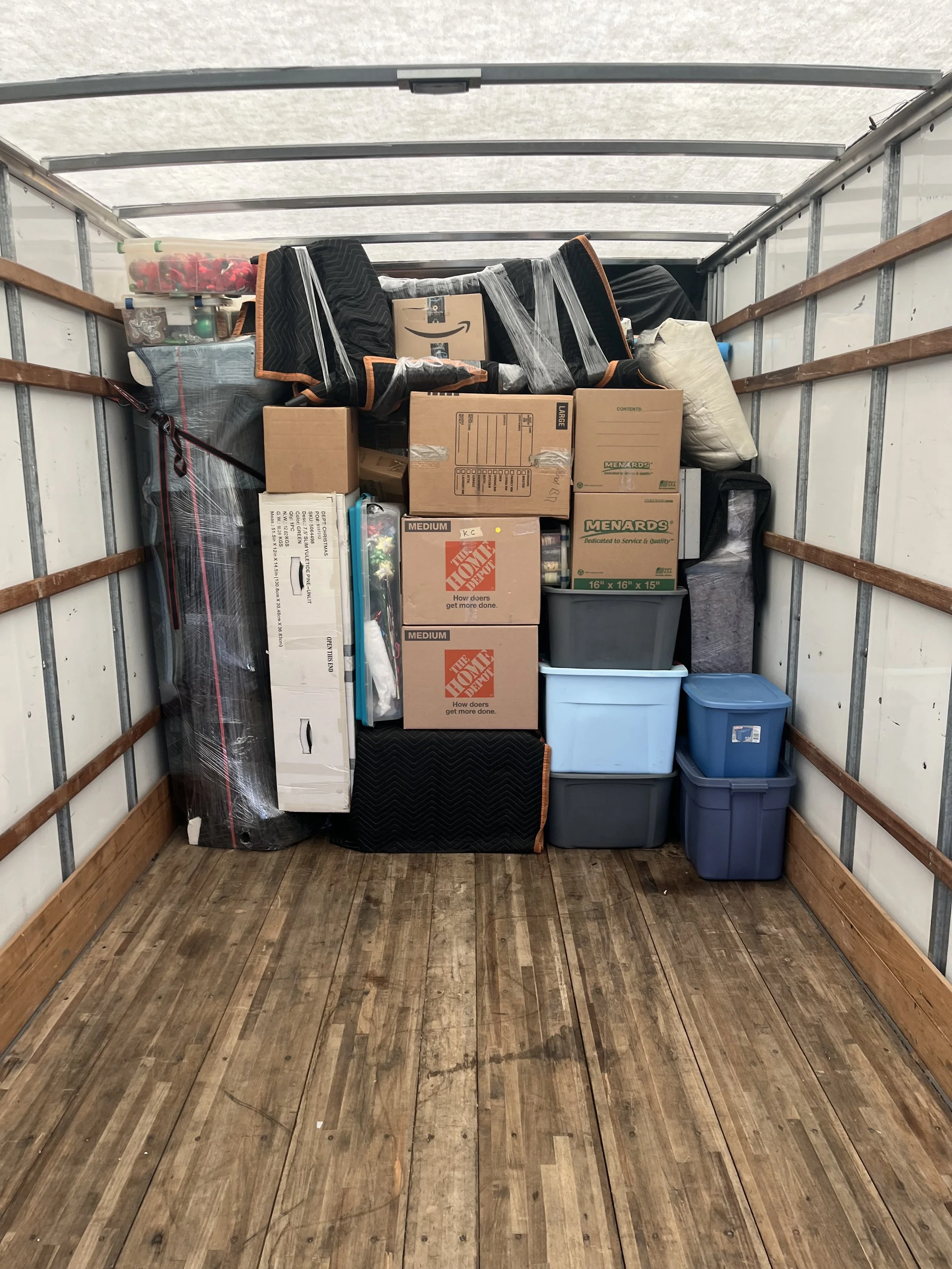 The inside of a storage container filled with boxes, plastic bins, and wrapped furniture on a wooden floor.