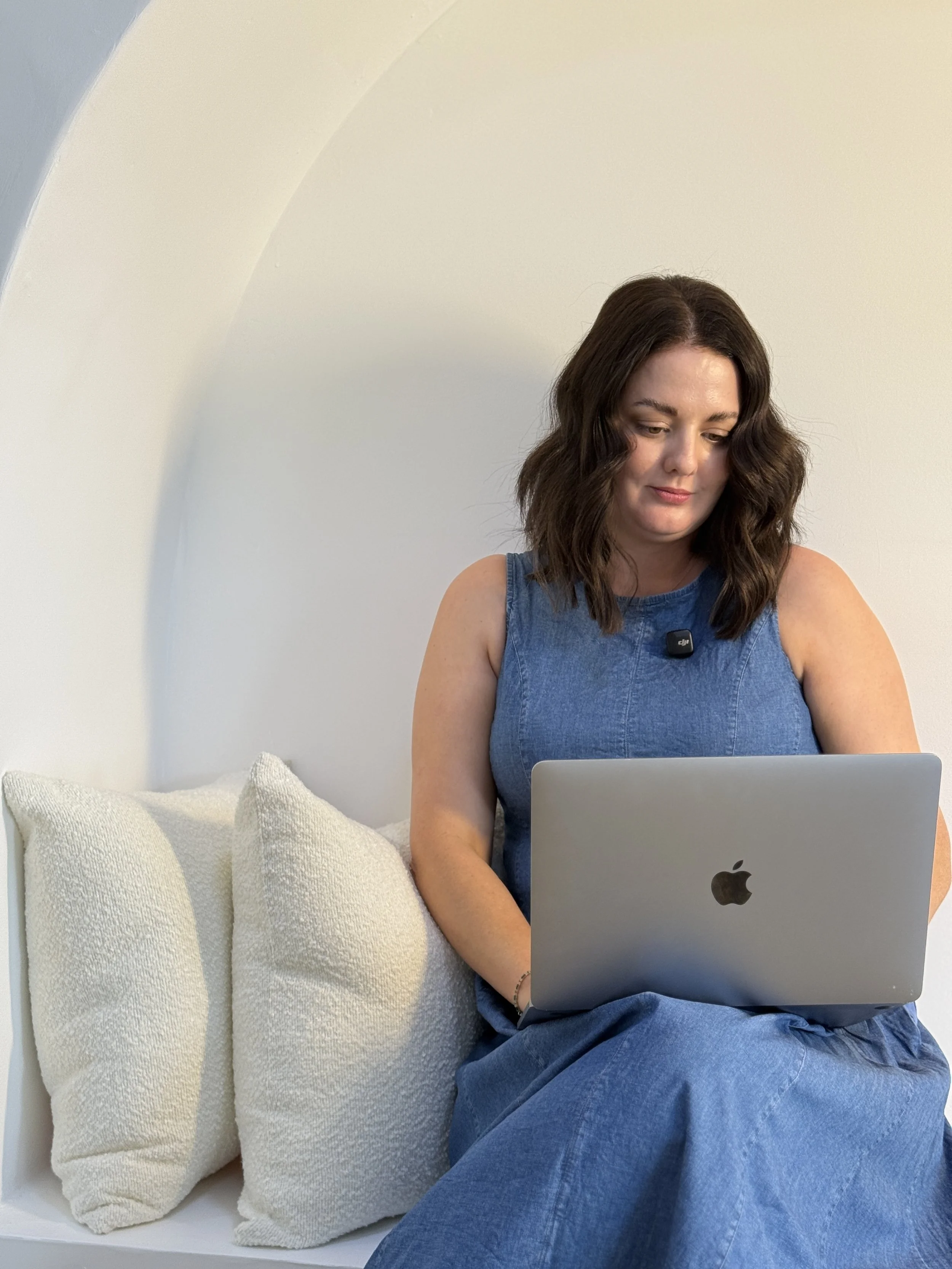 A woman sitting on a white bench with cream pillows, working on a silver MacBook. This is Holly Newman, owner of Lemon Creative, Social Media Manager, Content Creator in Townsville QLD