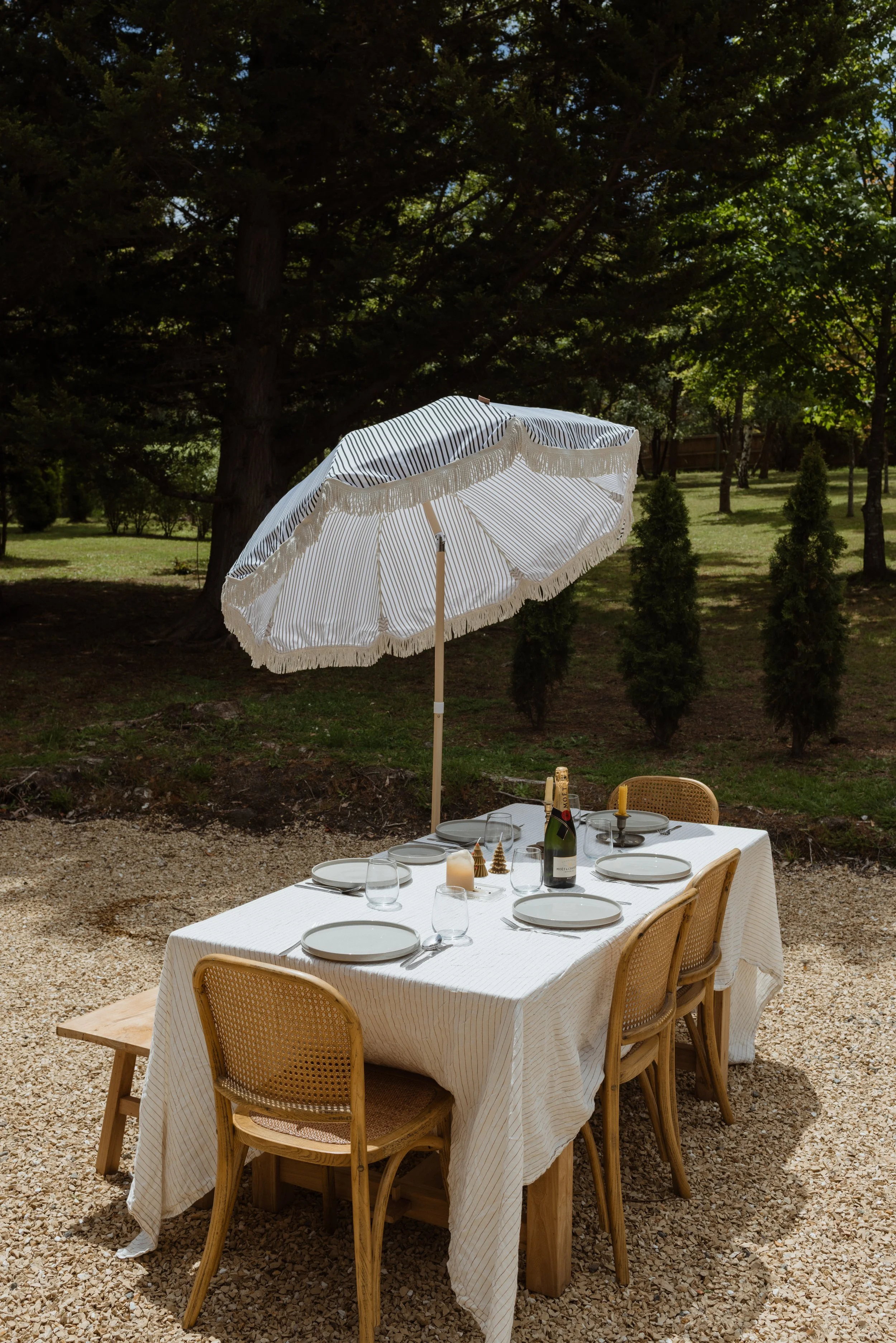 Outdoor dining table set for four with white tablecloth, plates, glasses, and a bottle of champagne under a striped umbrella in a garden setting with trees and shrubs in the background.