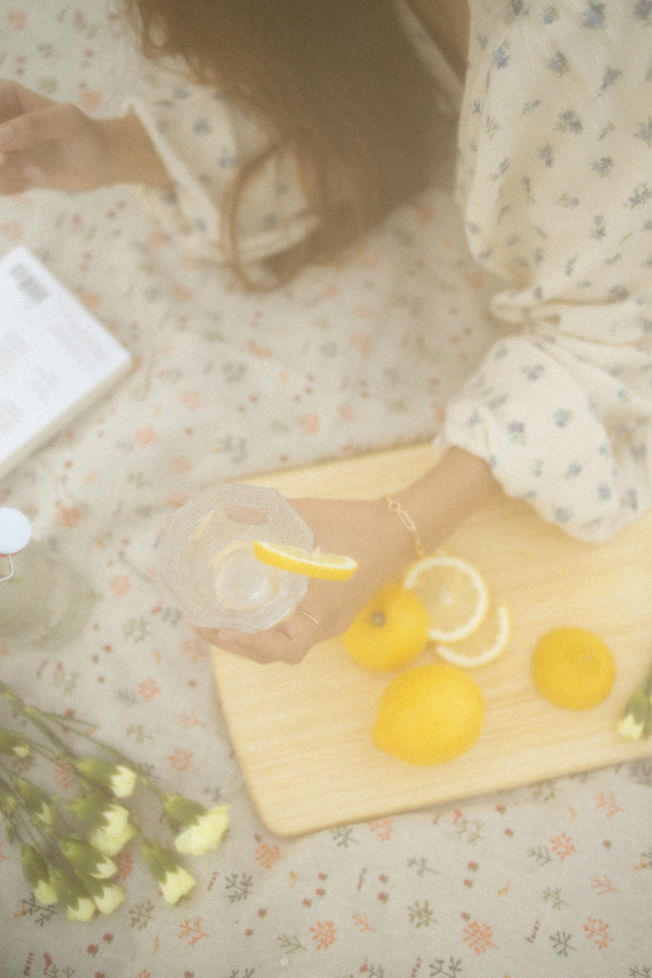 Person holding a glass of lemon water with yellow lemons and lemon slices on a wooden cutting board, floral tablecloth background.