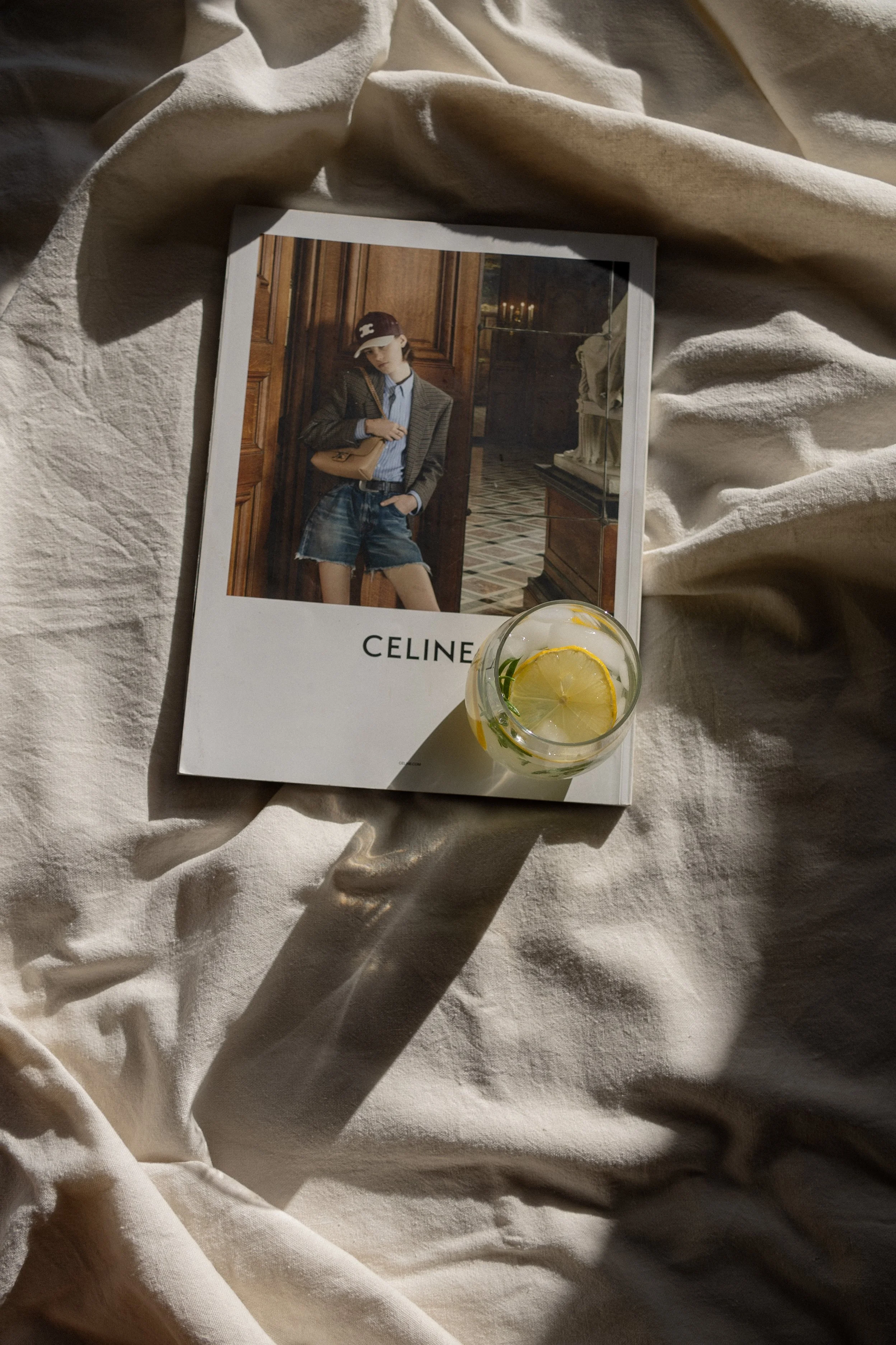A magazine featuring a woman in a blazer, shorts, and cap, with a glass of lemon water and a lemon slice, placed on a crumpled white fabric.