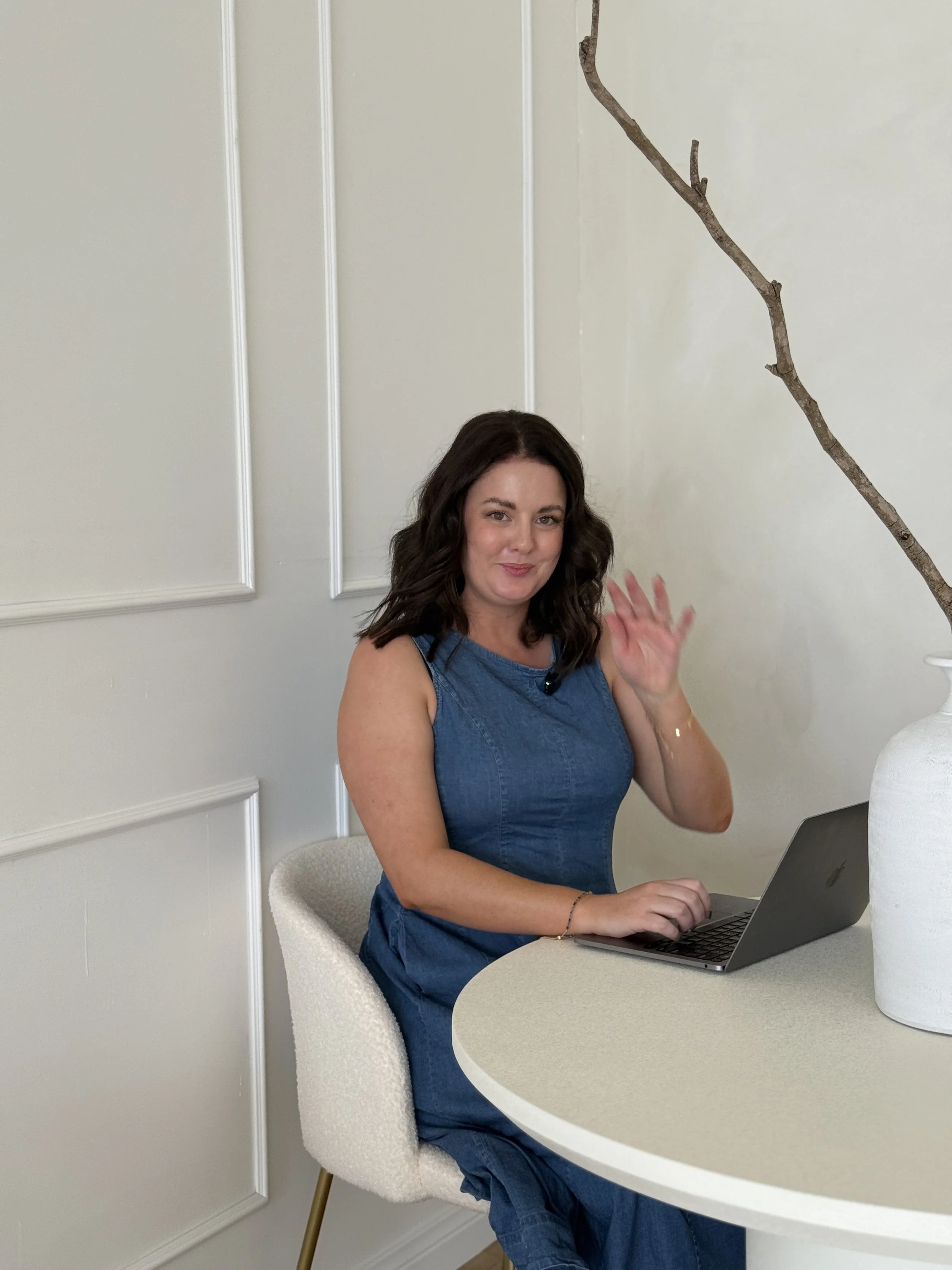 A woman with shoulder-length dark brown hair sitting at a round table with a laptop, waving and smiling, in a room with white walls and a decorative branch in a white vase. This is Holly Newman, owner of Lemon Creative