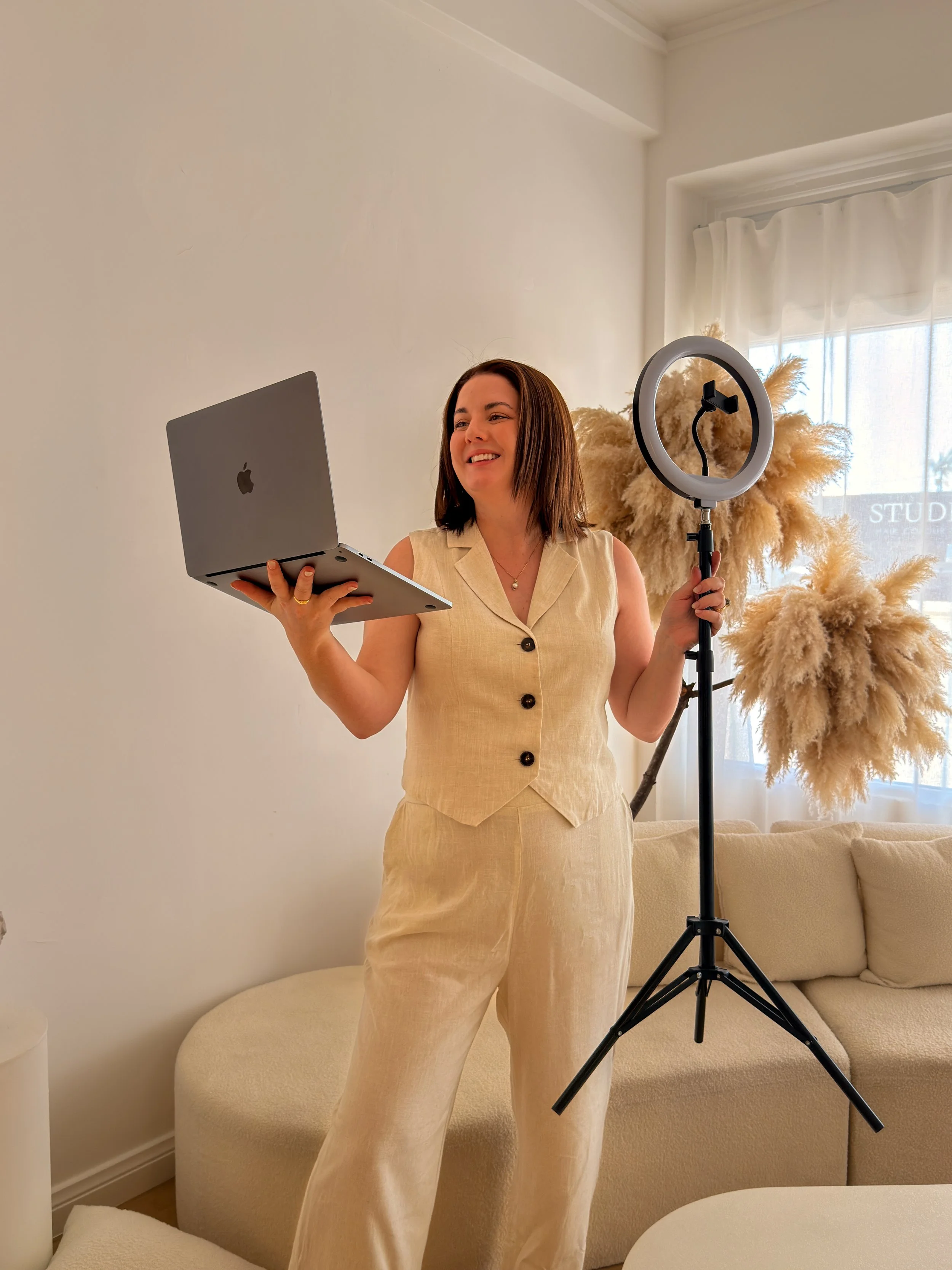 A woman in beige outfit holding a laptop and a ring light in a living room with a cream sofa and pampas grass decor. This is Holly Newman, owner of Lemon Creative, Social Media Manager, Content Creator in Townsville QLD