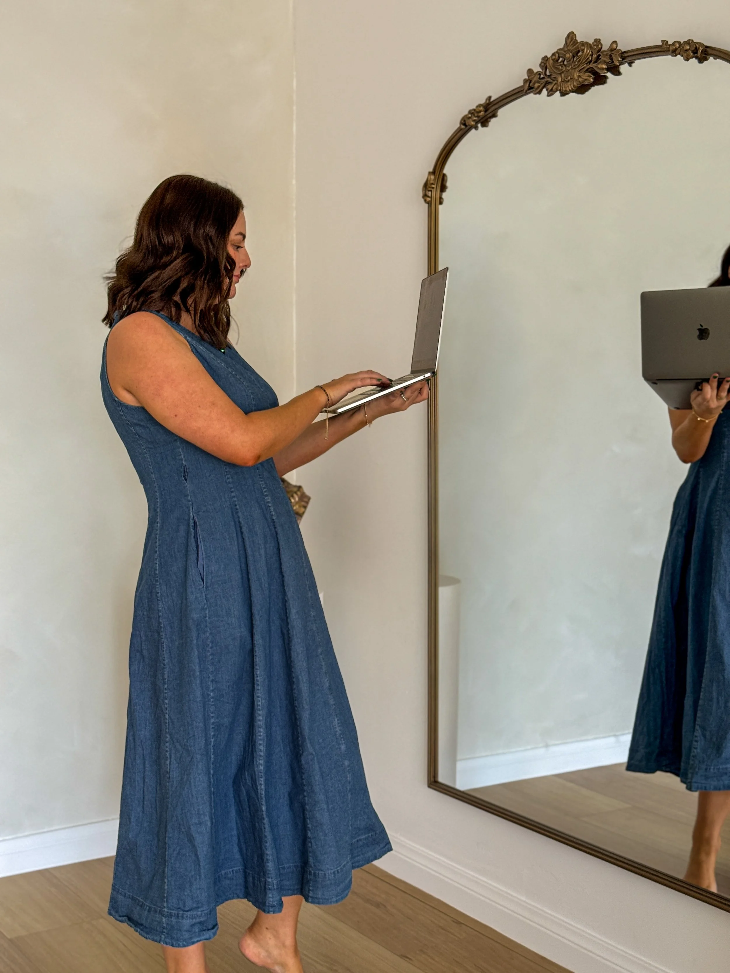 A woman in a denim dress taking a photo of her reflection in a large decorative mirror while holding a laptop. This is Holly Newman, owner of Lemon Creative, Social Media Manager, Content Creator in Townsville QLD
