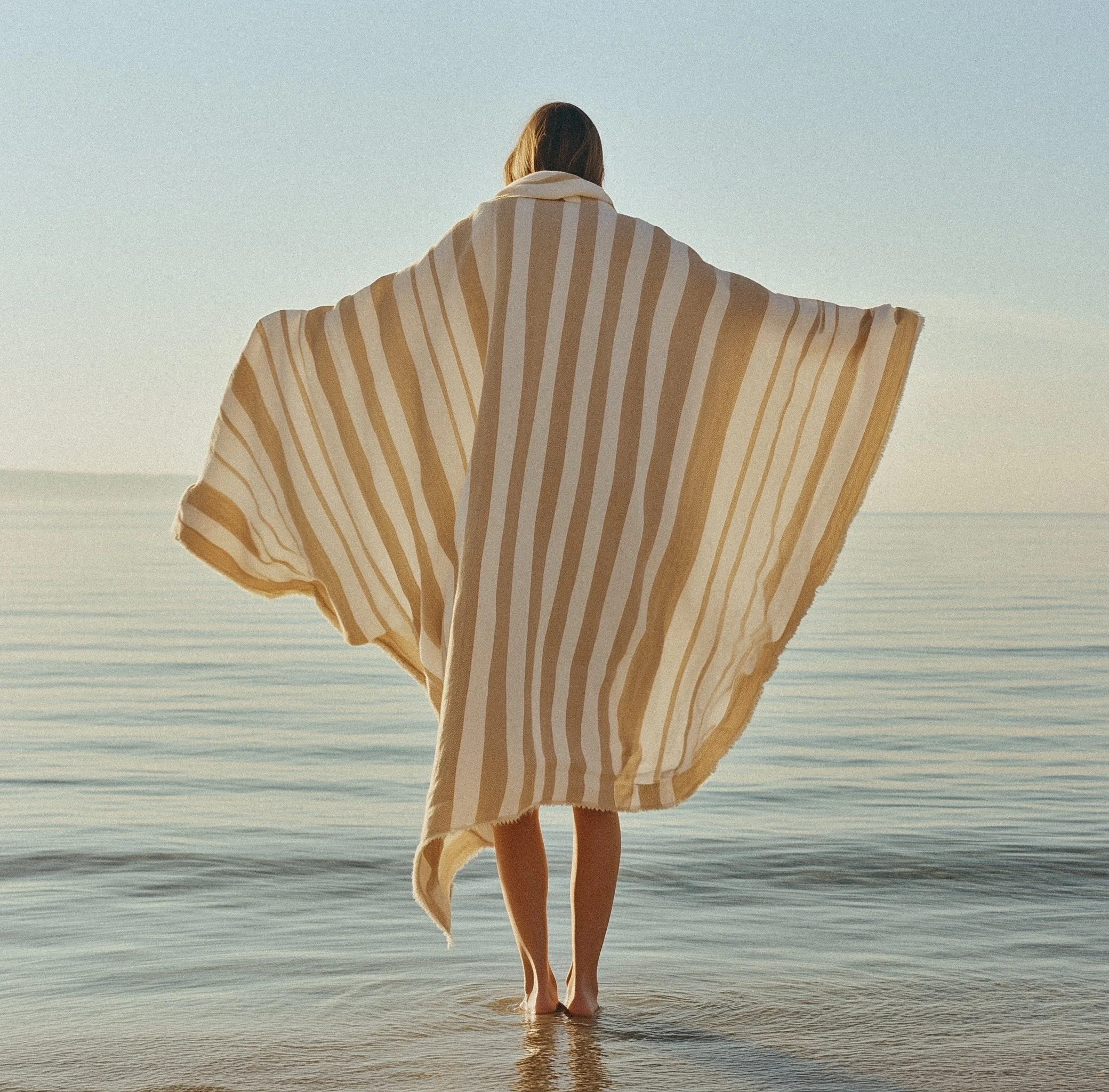 Person standing in shallow water at the beach, facing away, wrapped in a beige and white striped towel during sunset.