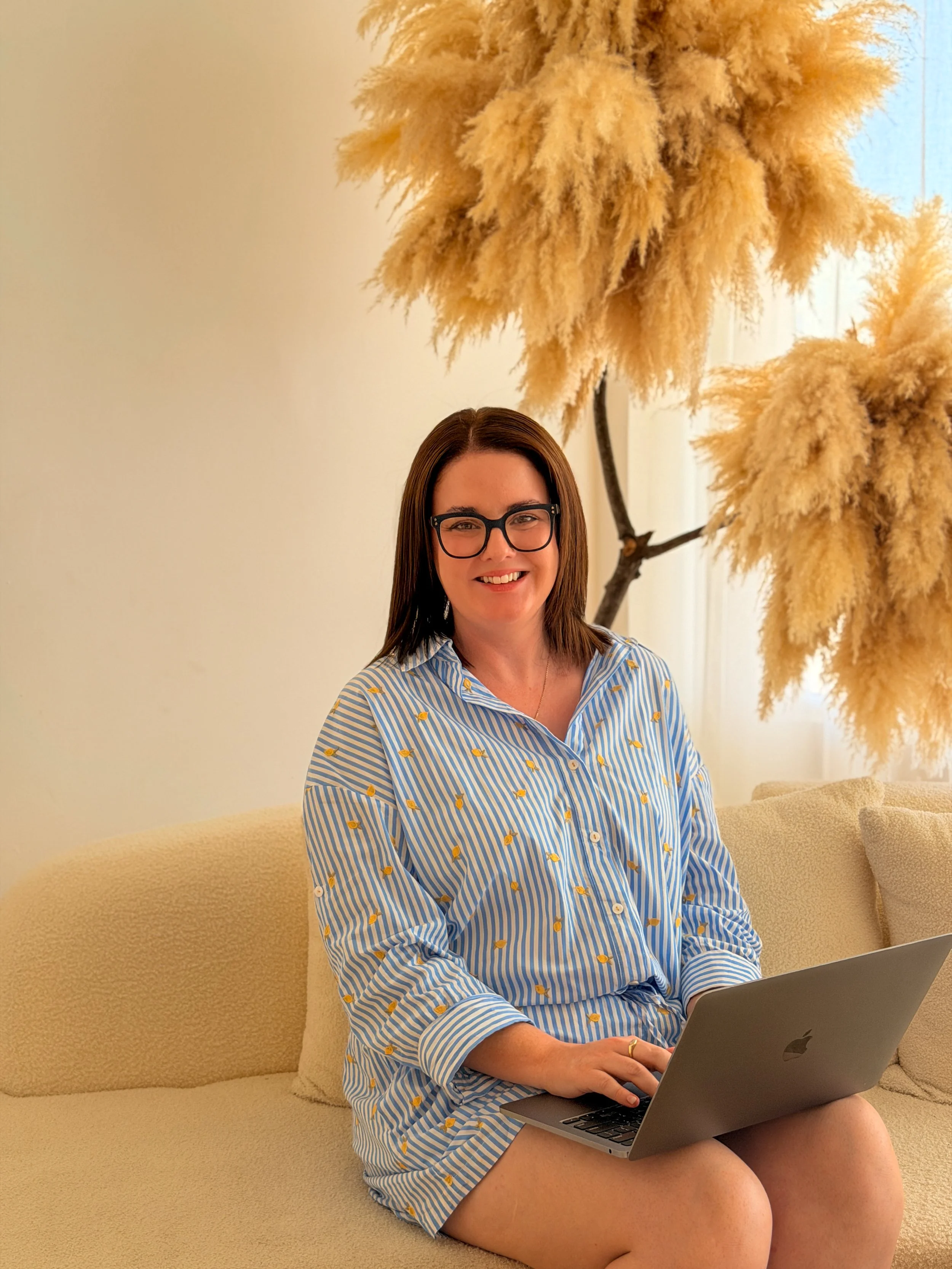 A woman with brown hair and glasses sitting on a beige sofa, smiling with a laptop on her lap, in a room with large pampas grass decor behind her and a window to the side. This is Holly Newman, owner of Lemon Creative, Social Media Manager