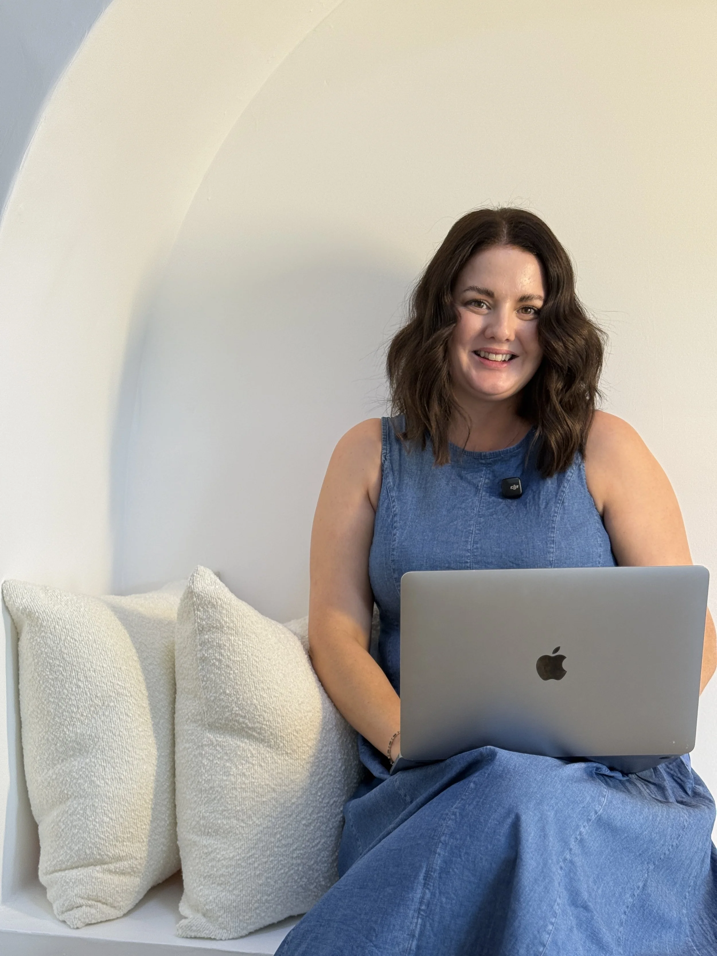 A woman with wavy brown hair wearing a denim dress, sitting on a white bench with cream-colored textured pillows, using a silver MacBook laptop. This is Holly Newman, owner of Lemon Creative, Social Media Manager, Content Creator in Townsville