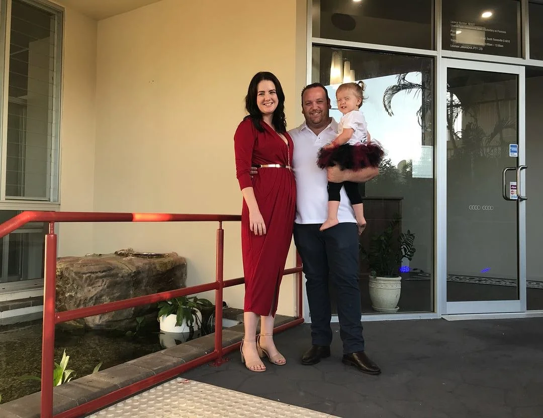 A smiling family of three standing outside a building. The mother is wearing a red dress, the father is in a white shirt and black pants, holding their young daughter dressed in a white top and black and red tutu. They are posing in front of glass doors with plants inside.