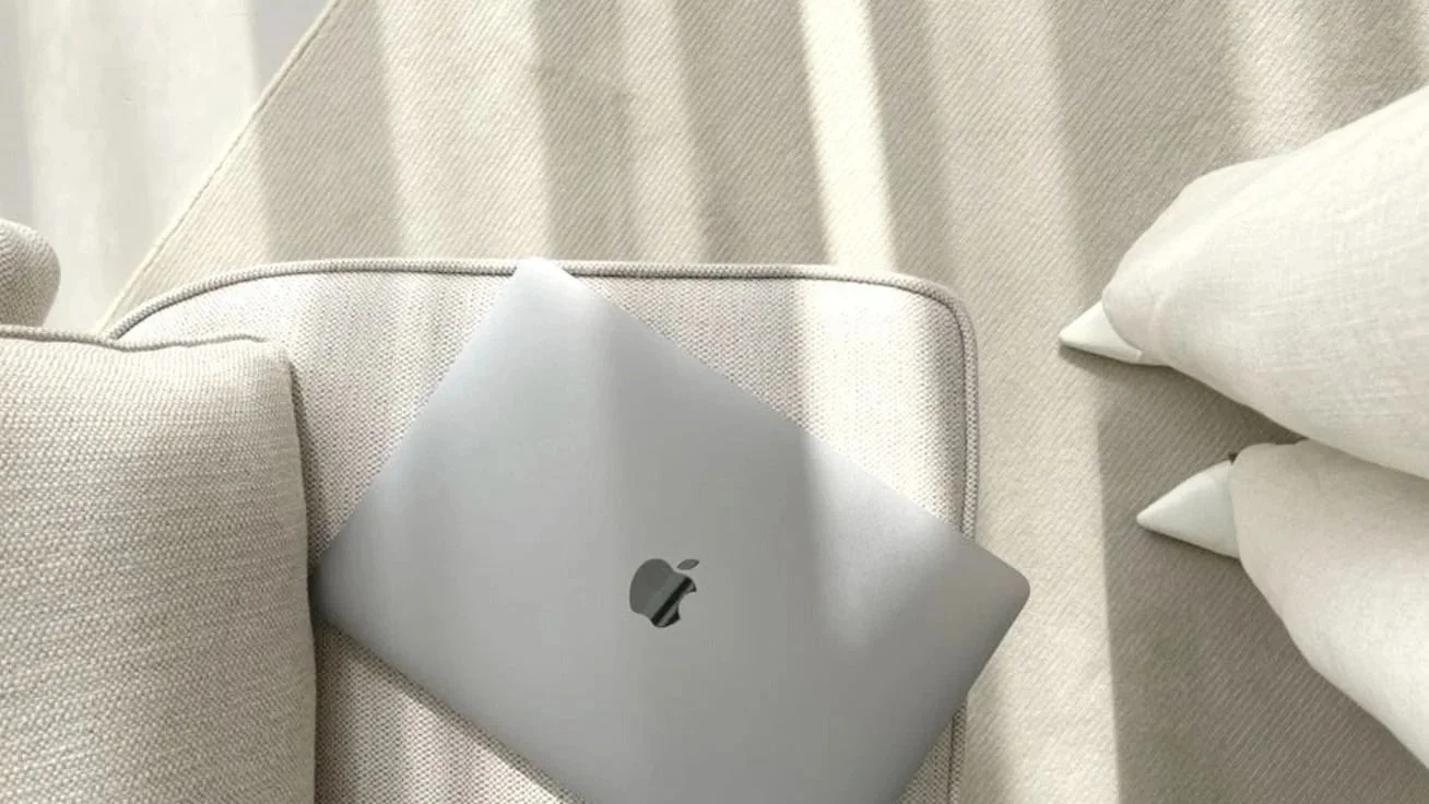 A silver Apple MacBook laptop on a beige couch with pillows, next to white cloud-textured curtains.