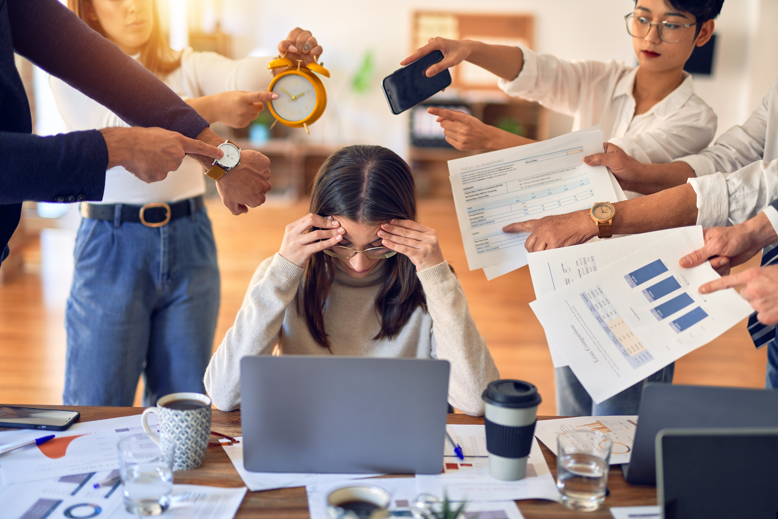 A woman with glasses sits at a cluttered desk with papers and coffee cups, holding her head in frustration while multiple people around her point and gesture urgently, indicating a stressful or chaotic work situation.