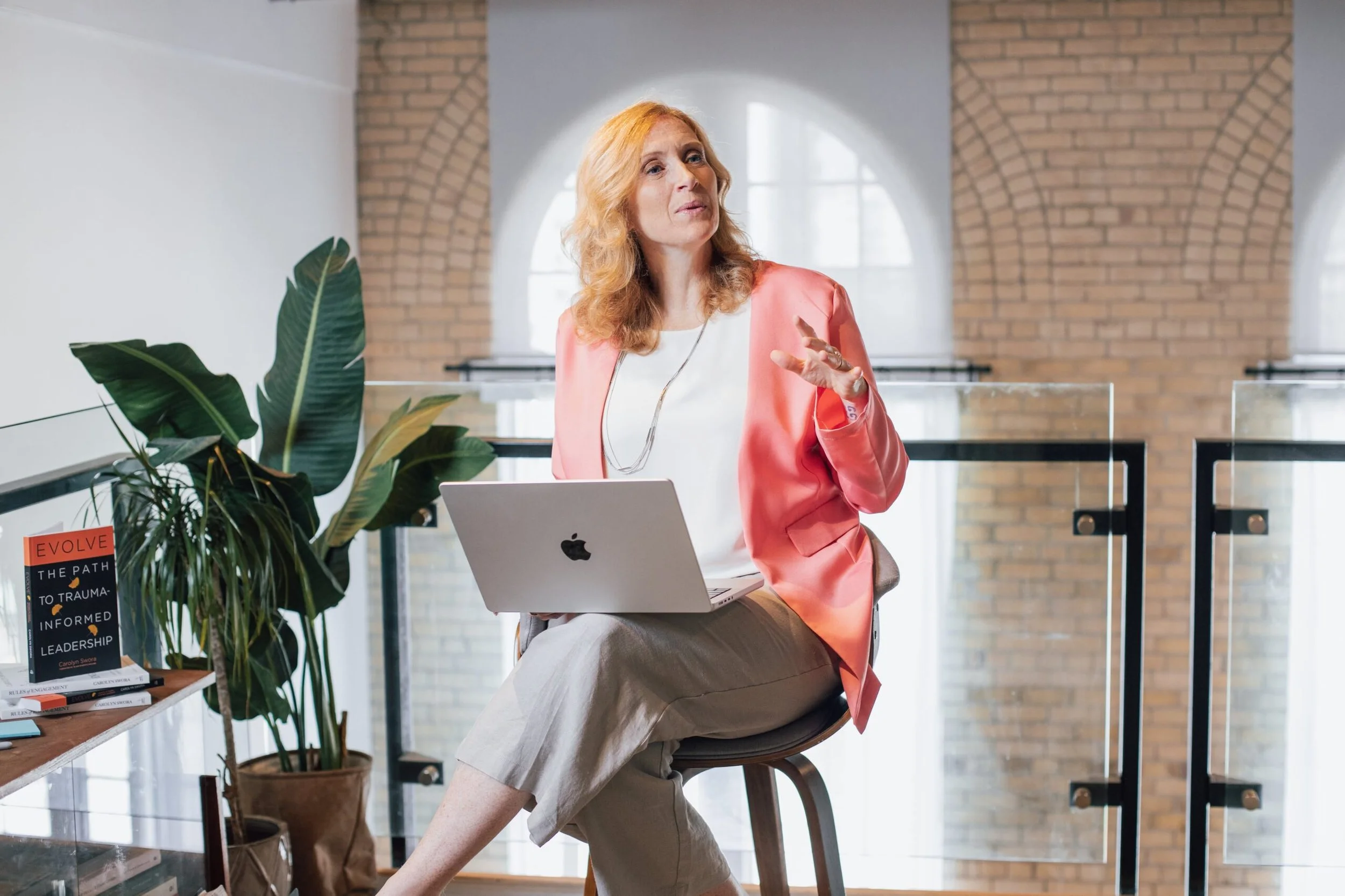 Carolyn Hart, co-founder of Resilient Voices, wearing a pink blazer and beige skirt, sitting on a bar stool in a modern office space, gesturing as she talks, with a laptop in front of her and books and plants nearby.