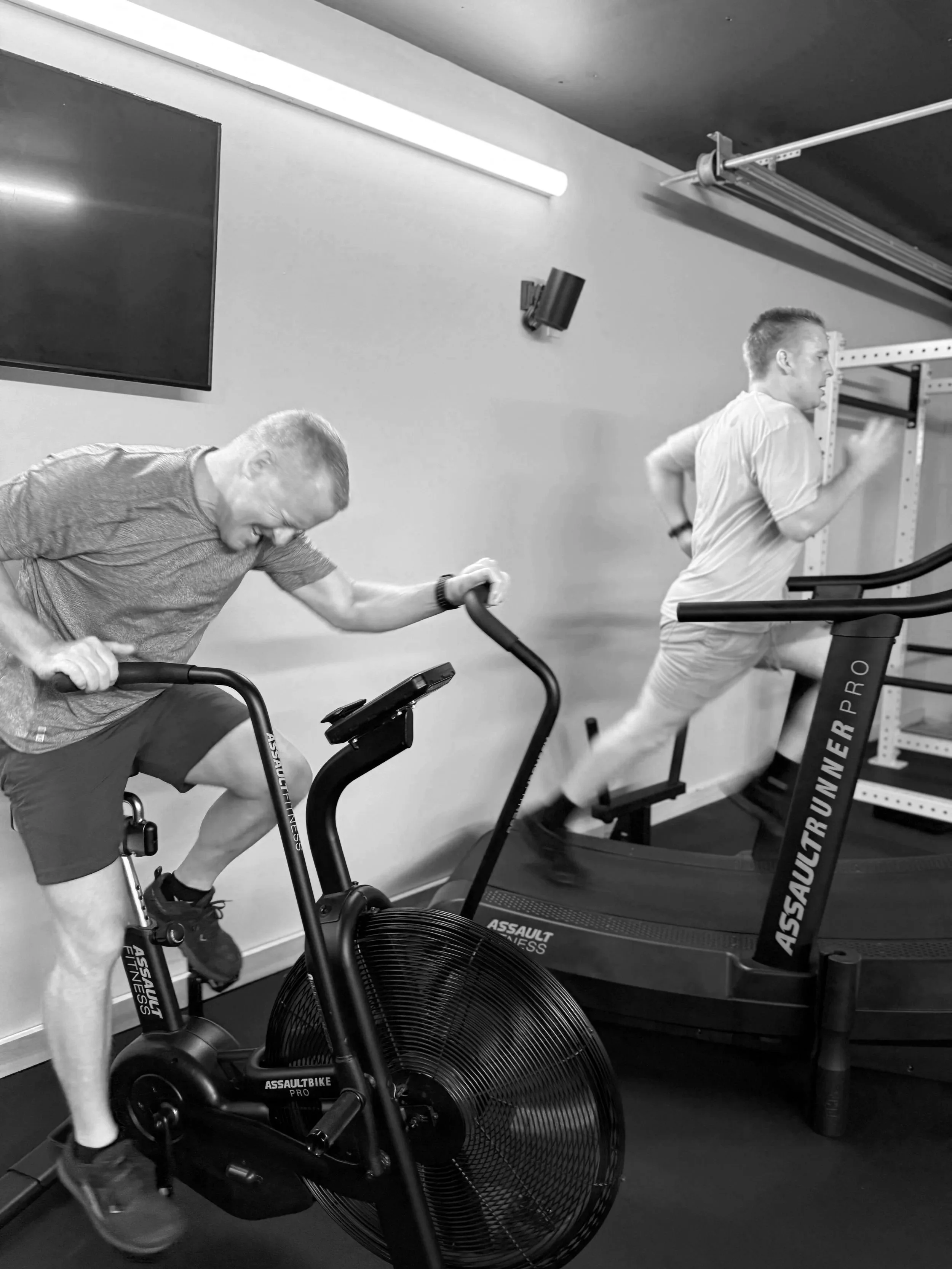 Two men working out on exercise equipment in a gym, one on a stationary bike and the other on a treadmill, in black and white.
