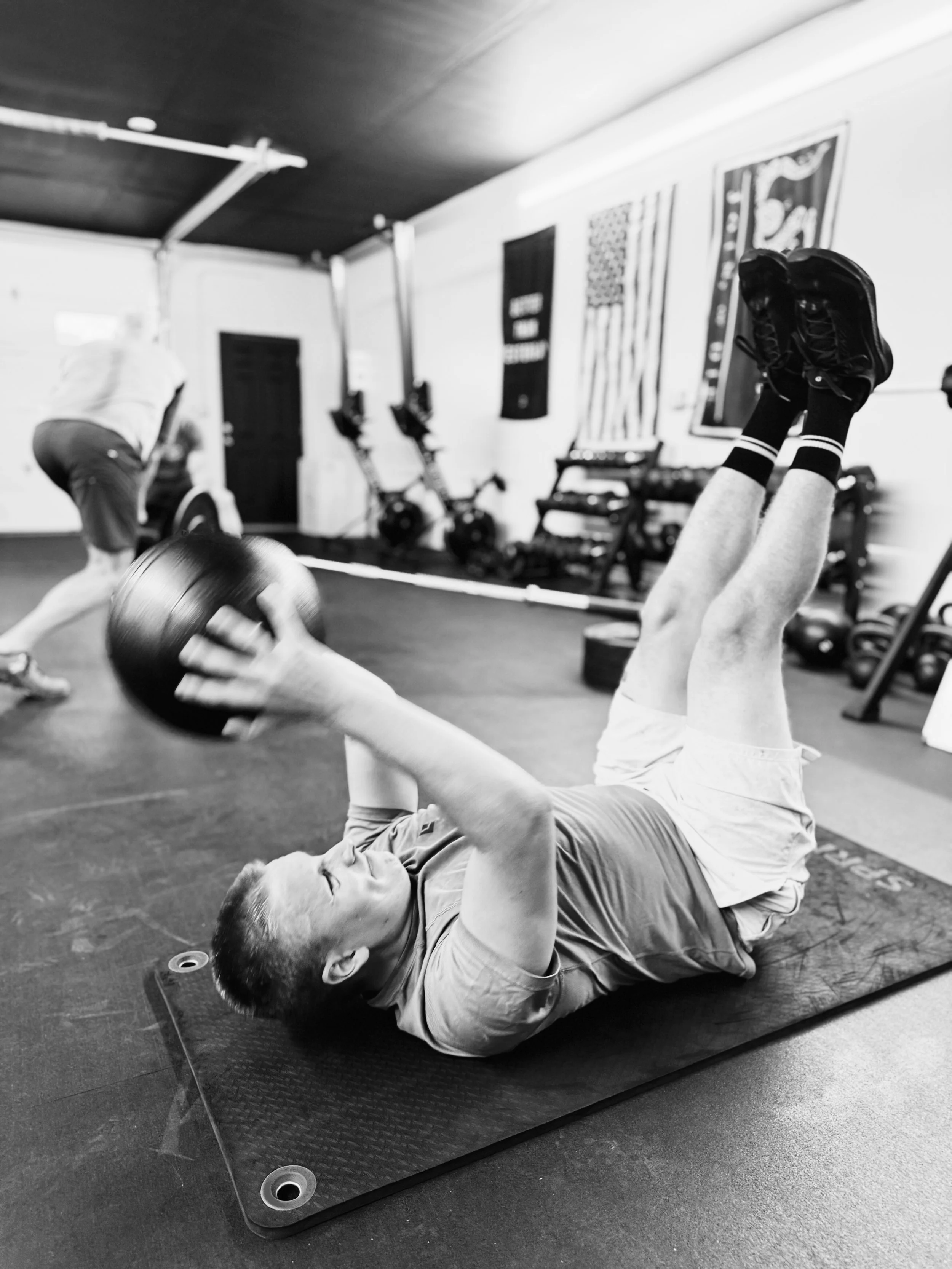 A young man doing a crunch exercise with a medicine ball in a gym, with other gym equipment and person in the background.