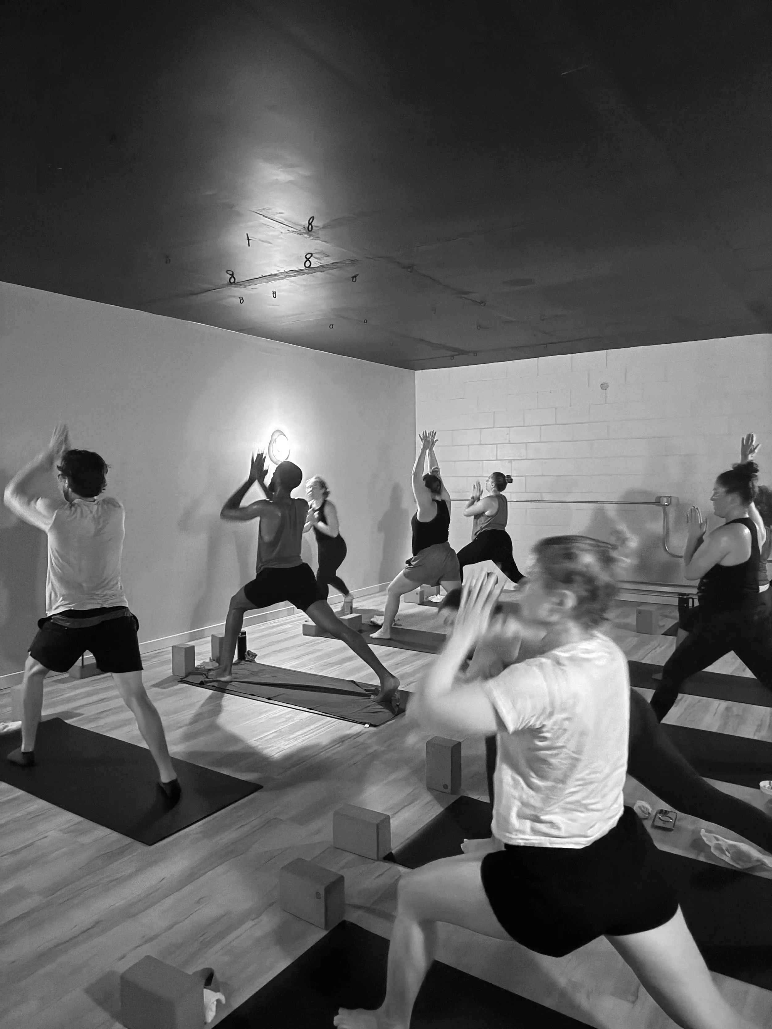 People participating in a yoga class, practicing poses on mats in a studio with wooden floors and plain walls.