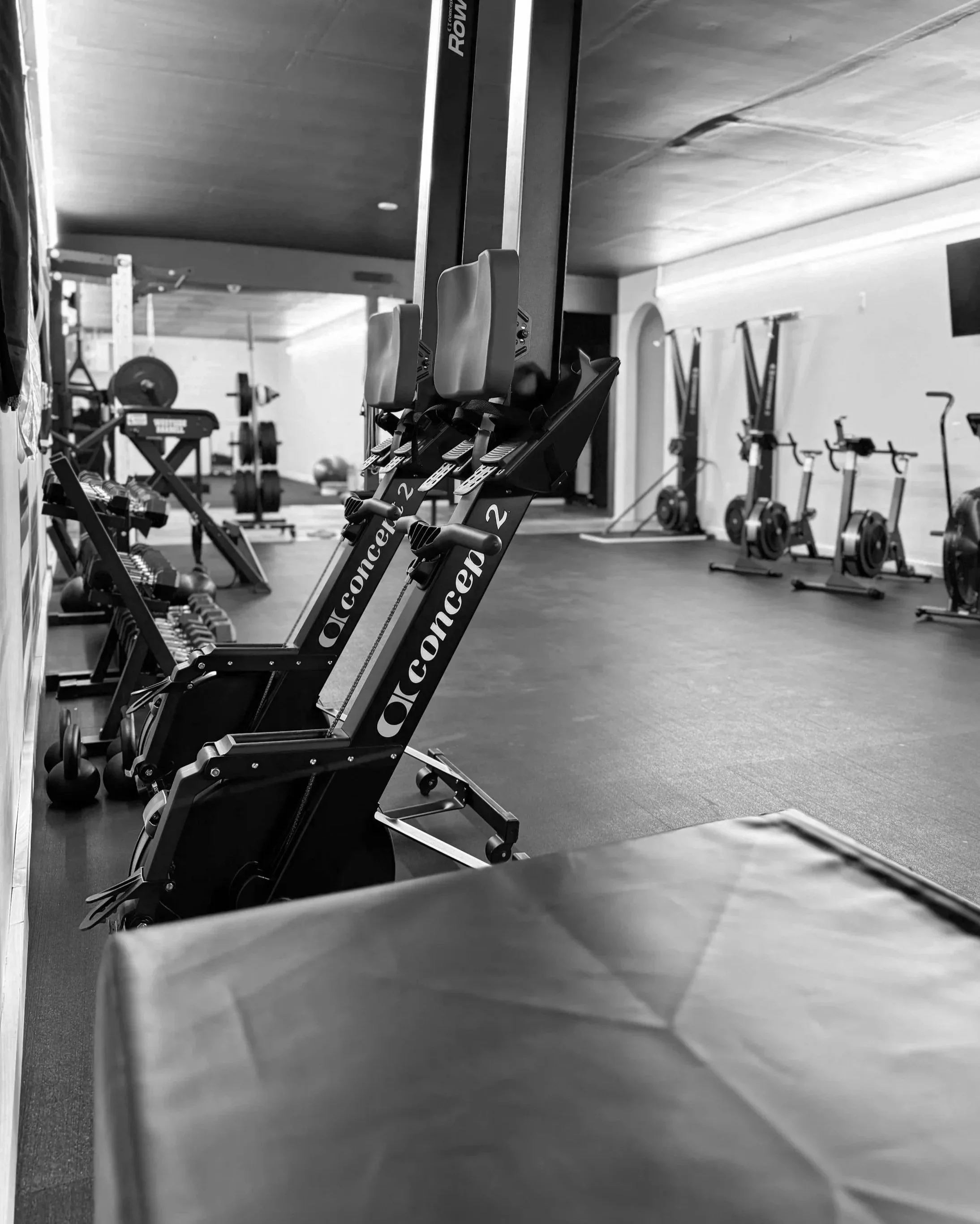 A black and white photo of a gym with exercise equipment, including indoor rowing machines in the foreground and stationary bikes in the background.