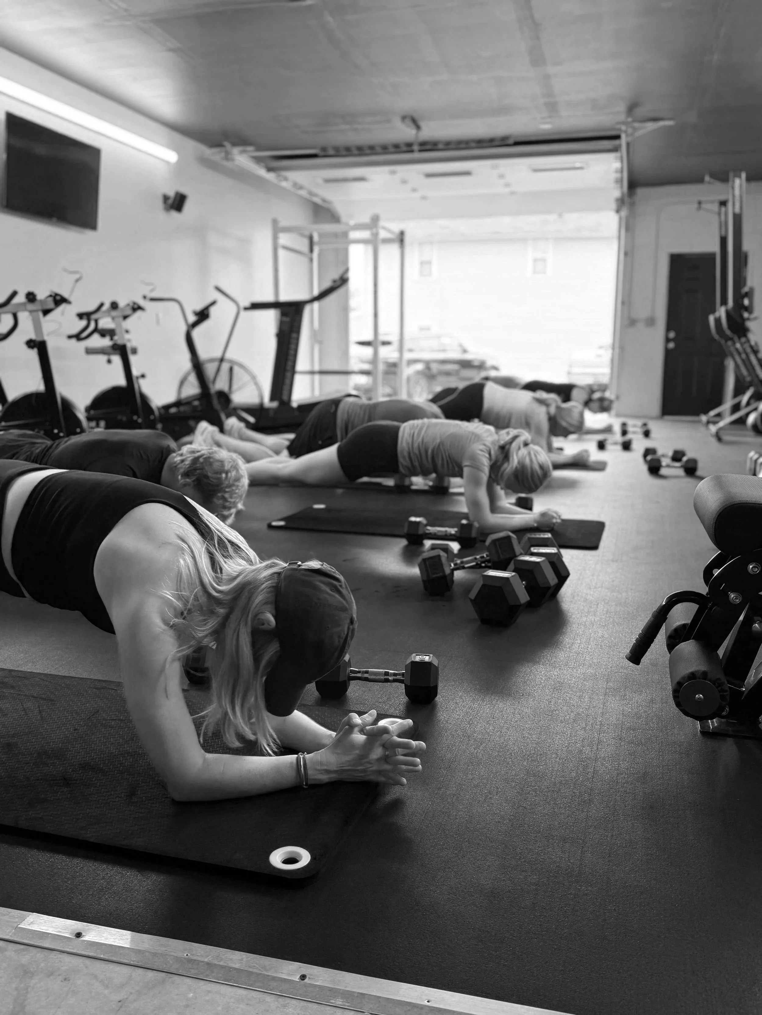 Group of people doing planks on yoga mats inside a gym, with dumbbells scattered around, in black and white.