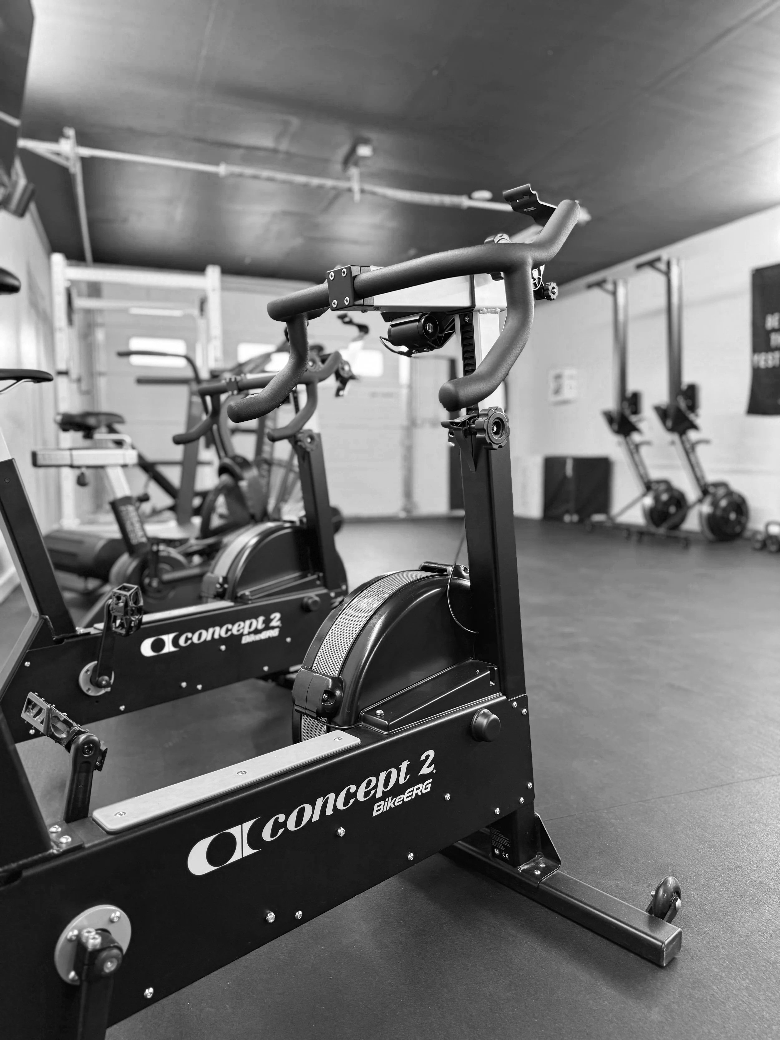 Indoor gym with exercise bikes, including a black Concept 2 BikeErg in the foreground, and other cardio equipment in the background, black and white photo.