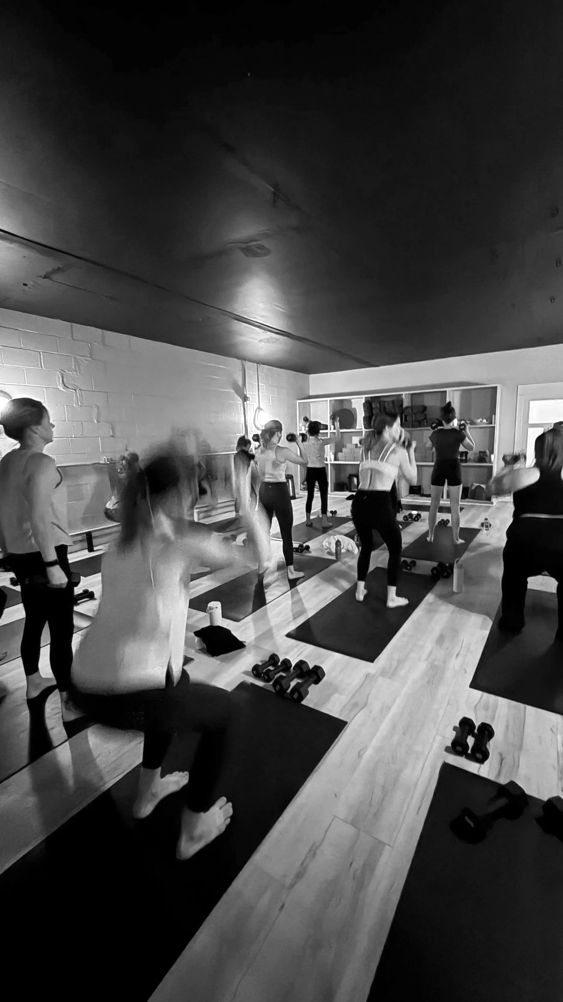 Women participating in a group fitness class or yoga session in a studio, standing on mats with dumbbells around them, in black and white.