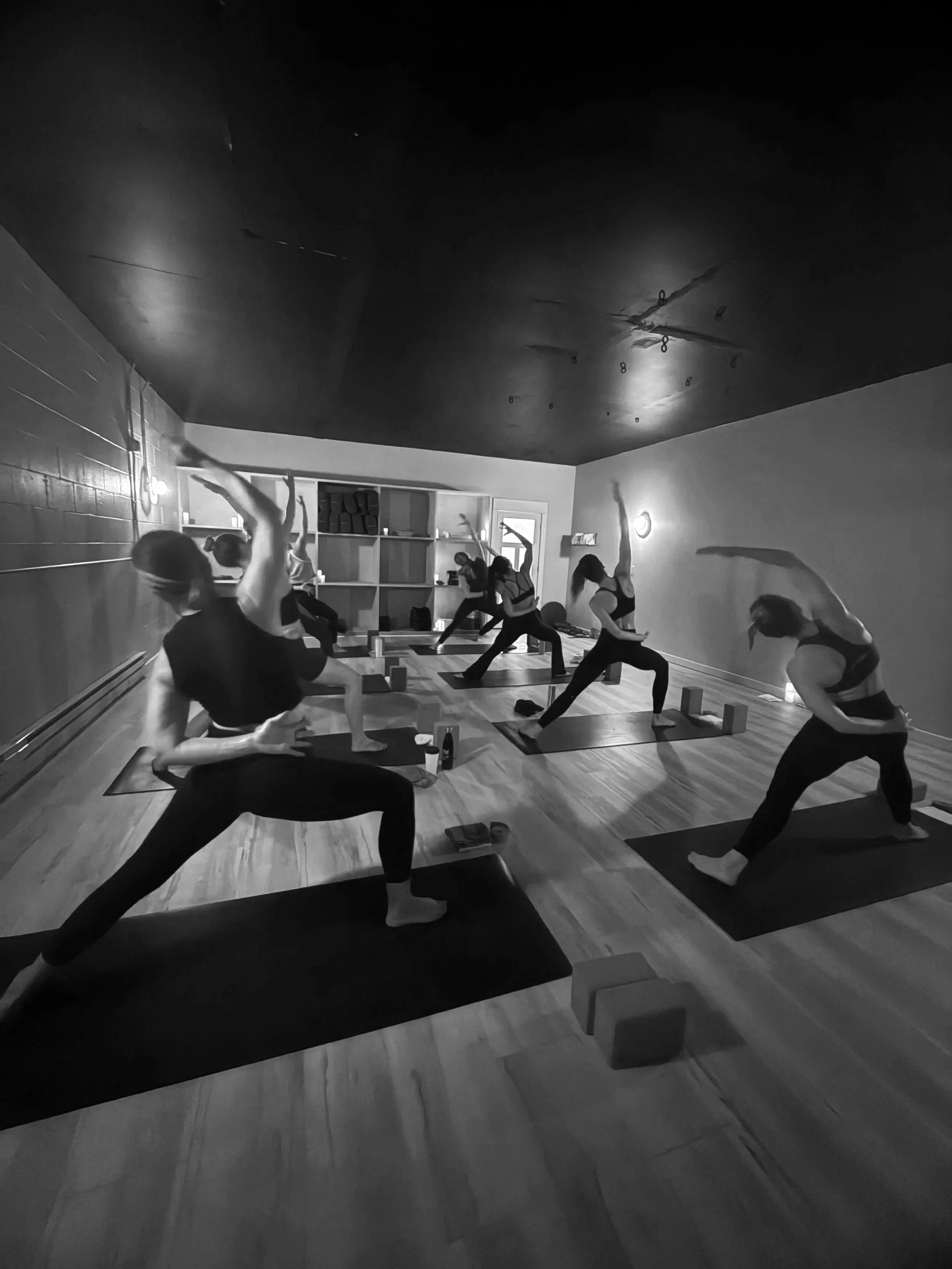 A group of women practicing yoga in a dimly lit studio, performing side stretches on mats, with yoga props and shelves in the background.