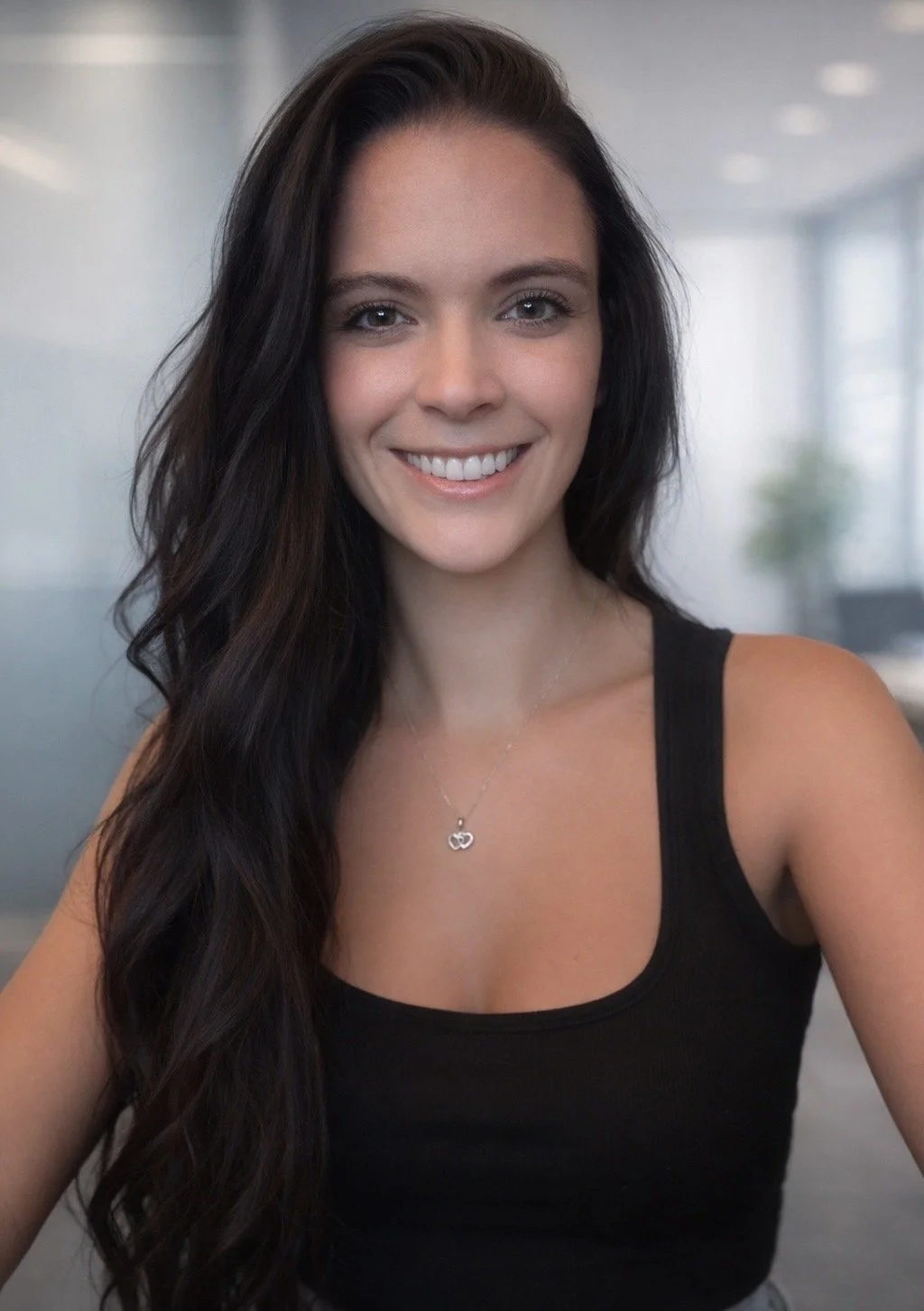 A woman with long dark hair smiling, wearing a black sleeveless top and a necklace with an infinity symbol pendant, in a modern office setting with blurred background.