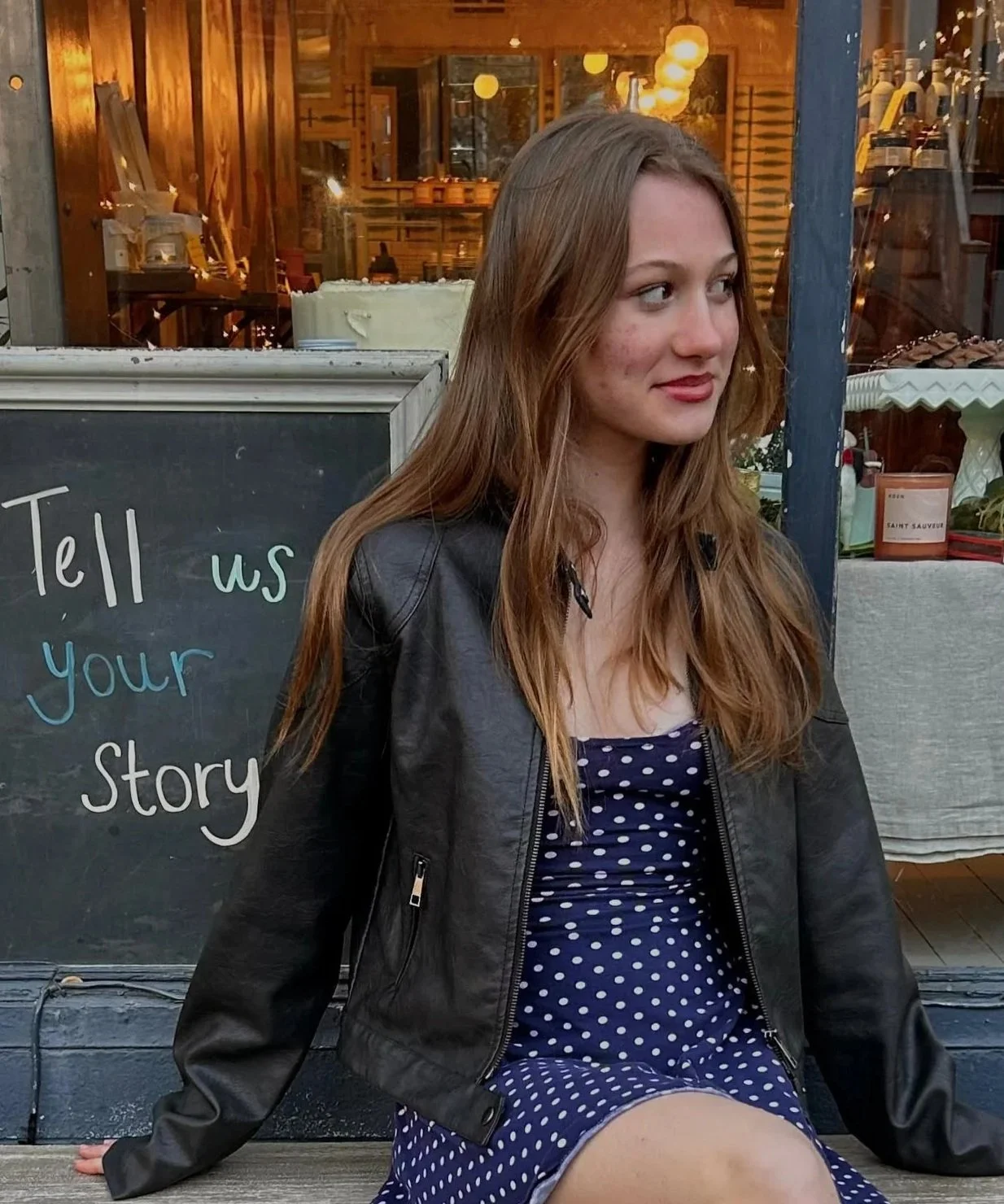 Young woman with long reddish hair sitting outside a cafe, wearing a navy polka dot dress and a black leather jacket, next to a blackboard sign that reads "Tell us your story."
