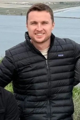 A man smiling outdoors near a body of water, wearing a black puffer jacket.