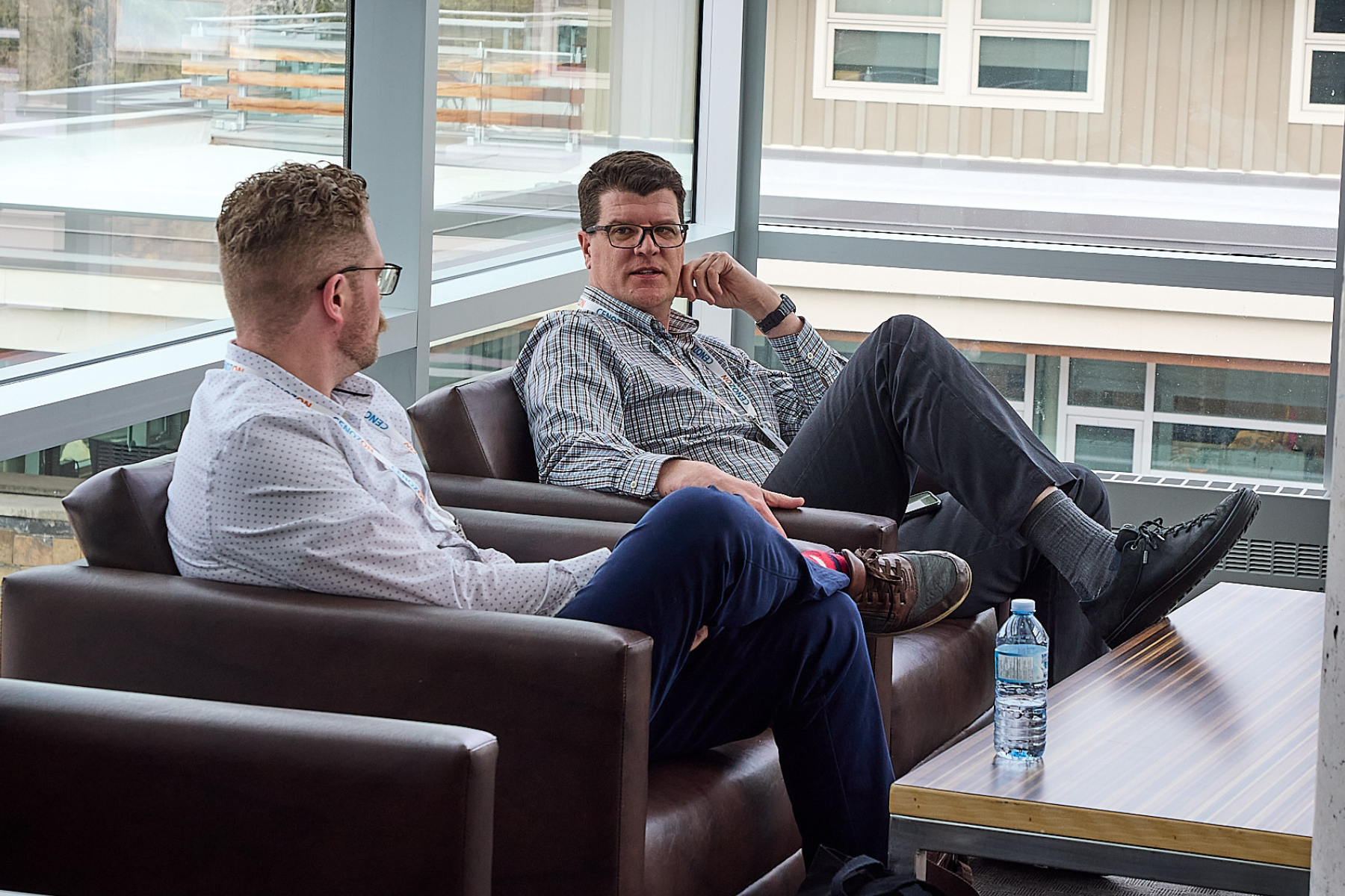 Two men sitting on brown sofas in a modern office or conference room, engaged in conversation. One man is wearing glasses and a checkered shirt, while the other is wearing a white, dotted shirt. A water bottle is on the table near them, and large windows with a view of a building's exterior provide natural light.