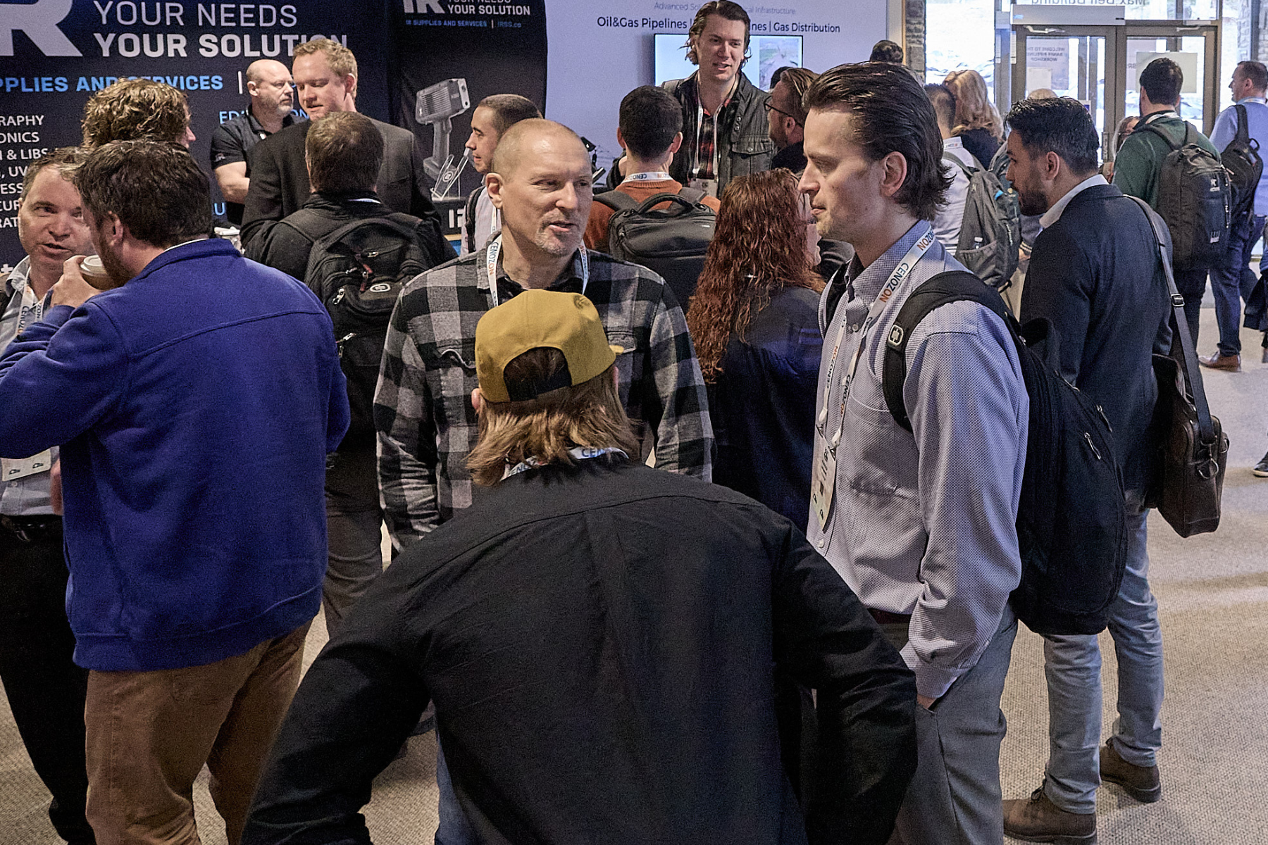 A group of people at a trade show or conference engaging in conversations, some wearing conference badges, with a backdrop featuring text related to oil, gas pipelines, and solutions.