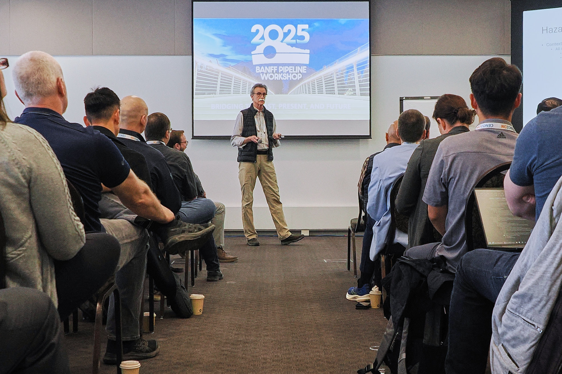 A man giving a presentation at the Banff Pipeline Workshop 2025, with a large screen behind him displaying the event logo and the theme 'Bridging the Past, Present, and Future.' Audience members are seated, listening attentively.