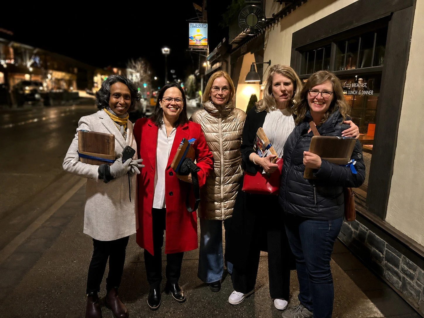 Five women standing together outside at night, smiling, holding wooden trays and folders, in front of a restaurant window with a sign that reads 'Daily Breakfast, Lunch & Dinner.'