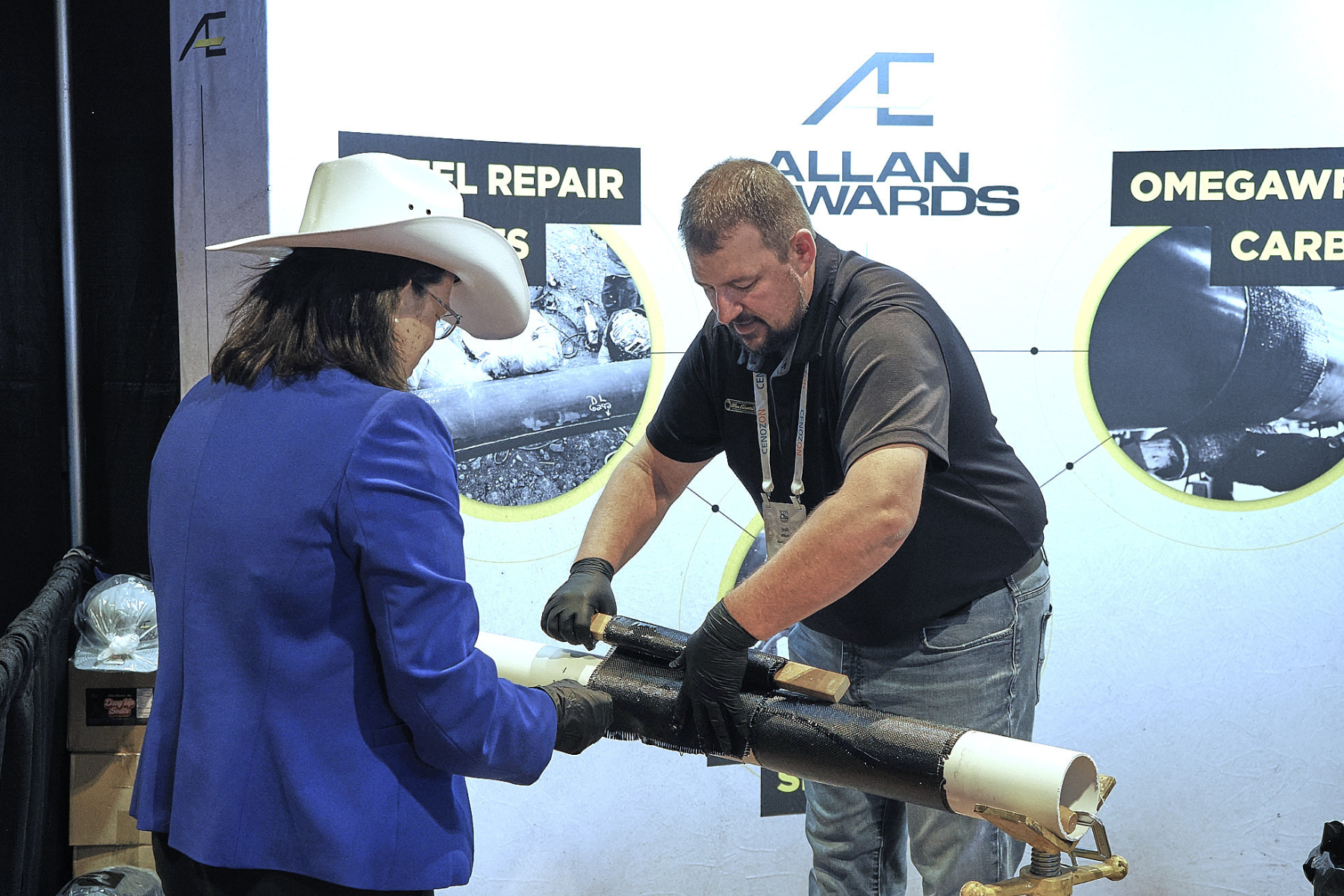 Two individuals working together on a project at a trade show, with displays behind them mentioning 'ALLAN WARDS', 'Omegwash', and 'Carp'. The person on the left is wearing a white cowboy hat and a blue blazer, while the person on the right is in a black shirt and gloves. They are handling a long cylindrical object, possibly a pipe, which is mounted on a worktable.