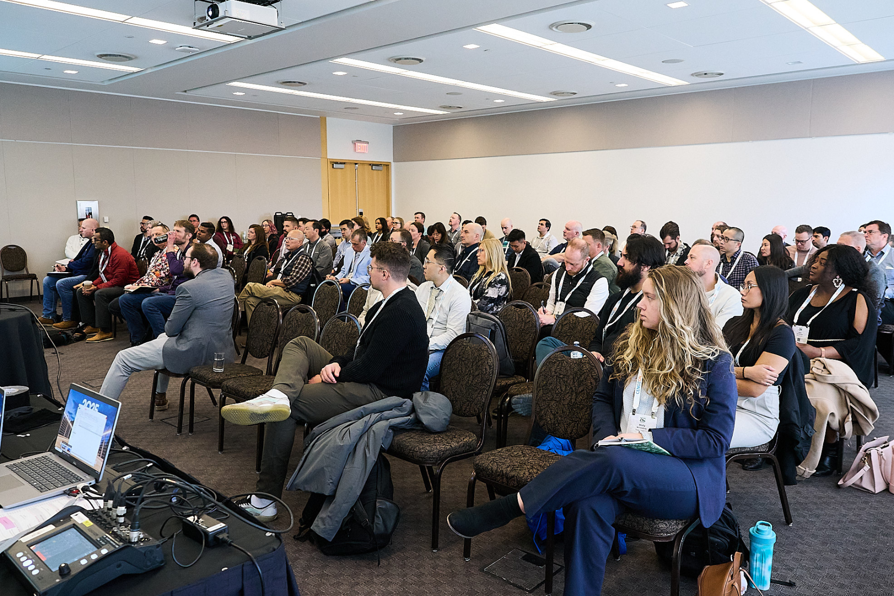 An audience of diverse people seated in rows, attending a conference in a large room with white walls and ceiling lights.