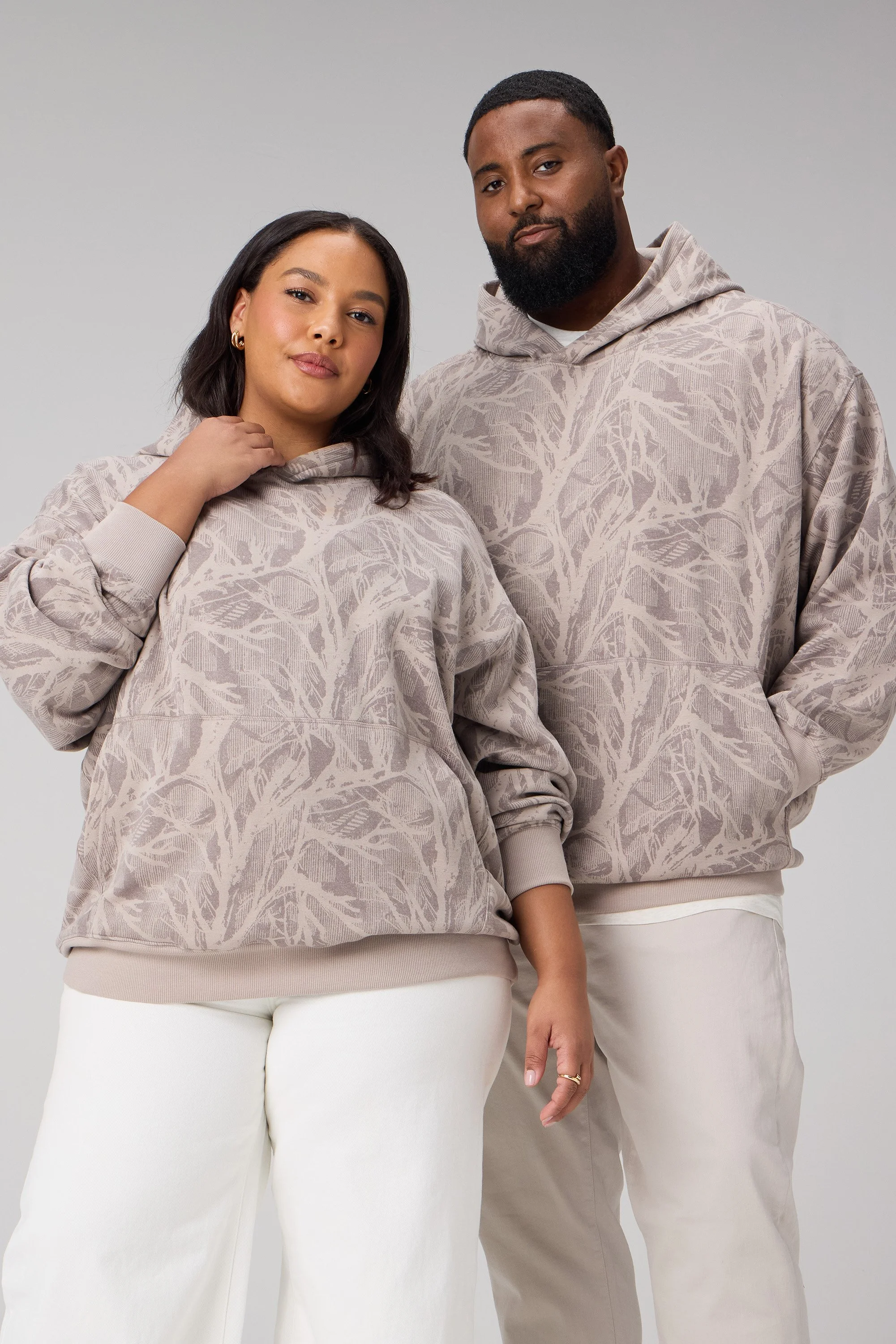 A woman and a man standing side by side wearing matching beige hoodies with a tree branch pattern, against a plain gray background.