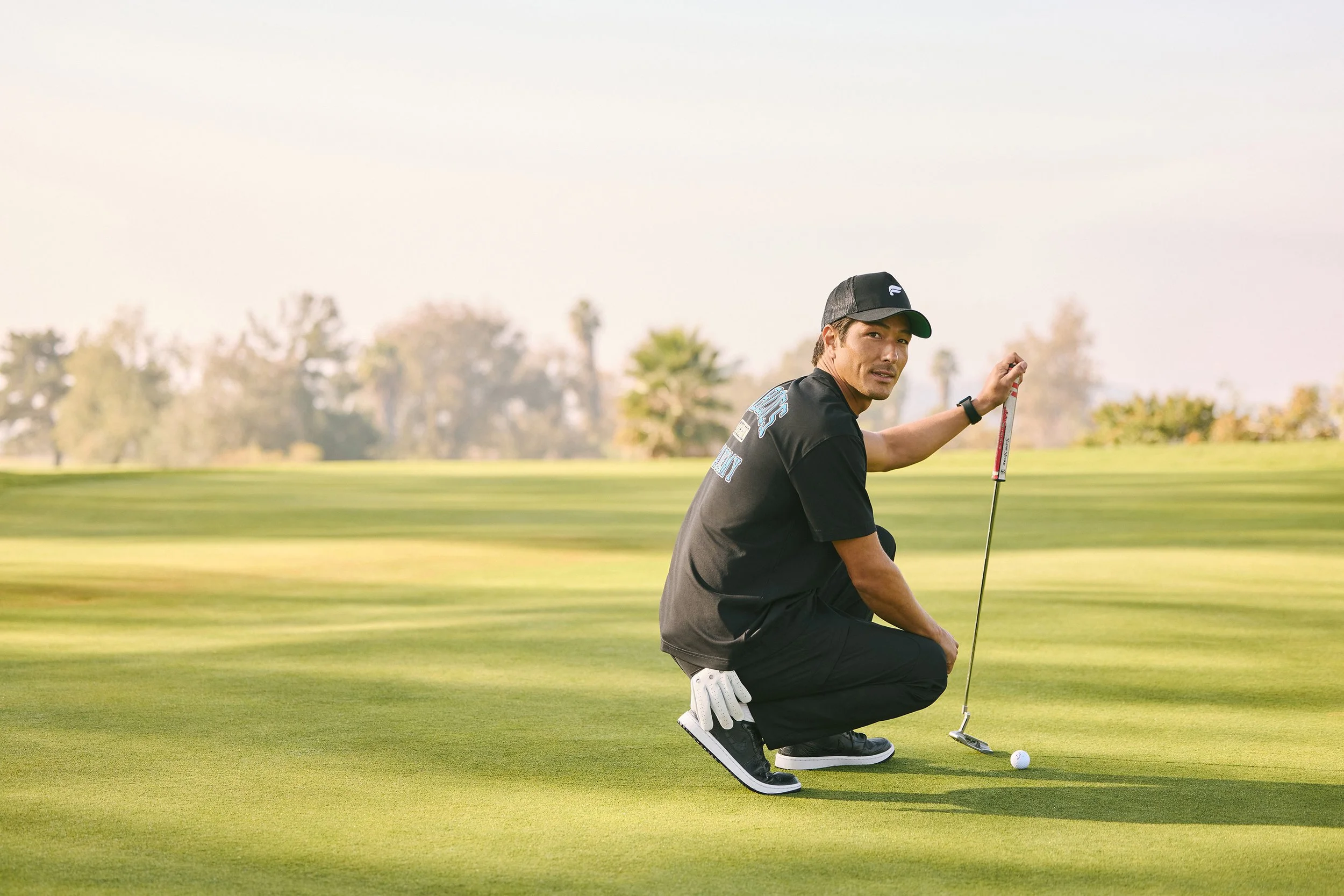 Art director on-figure e-commerce shoot, Los Angeles. A man crouching on a golf course with a golf club, preparing to hit a golf ball, with trees in the background.