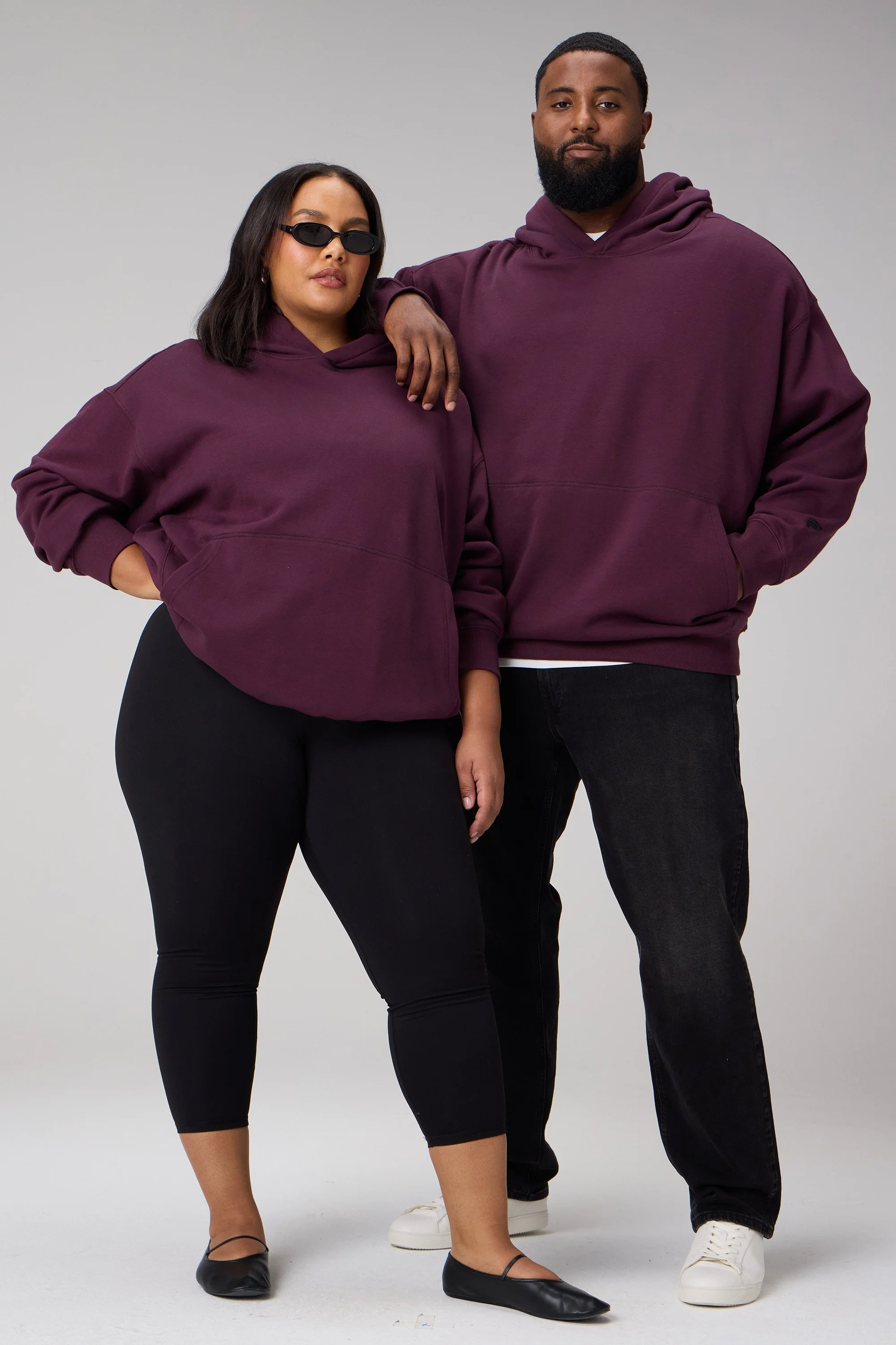 Two people, a woman and a man, stand side by side against a plain backdrop, both wearing matching dark purple hoodies and black pants, with the woman also wearing sunglasses.