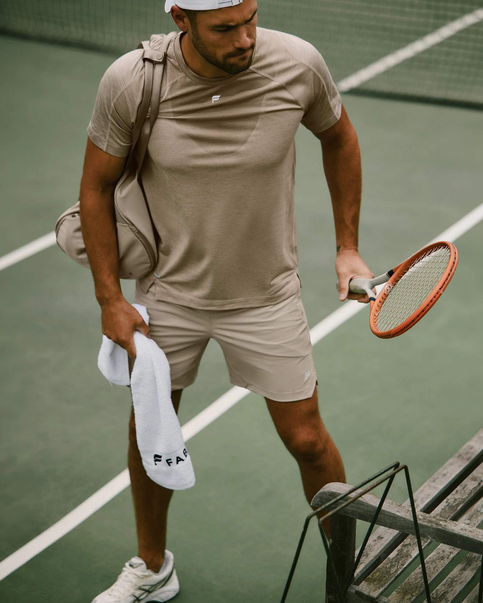 A man dressed in beige athletic clothing holding a tennis racket and towel on a tennis court.