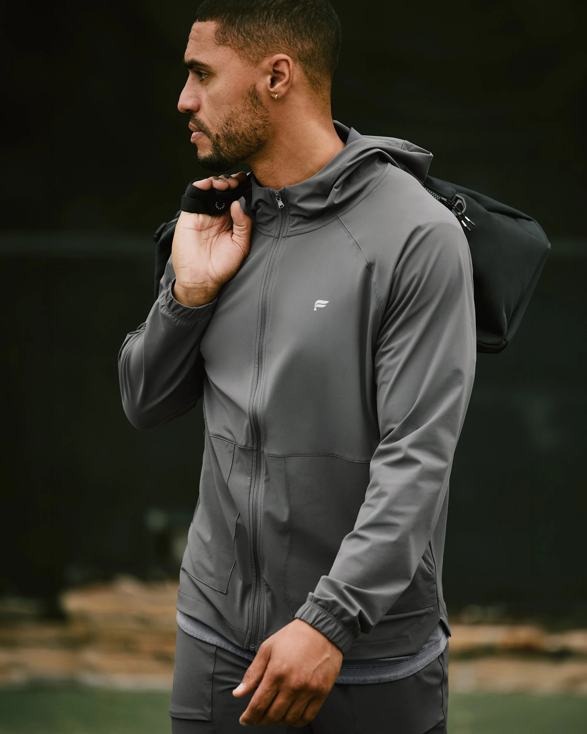 Art director on-figure e-commerce shoot, Los Angeles. Man wearing a gray athletic jacket and carrying a black backpack, holding a strap over his shoulder, outdoors.