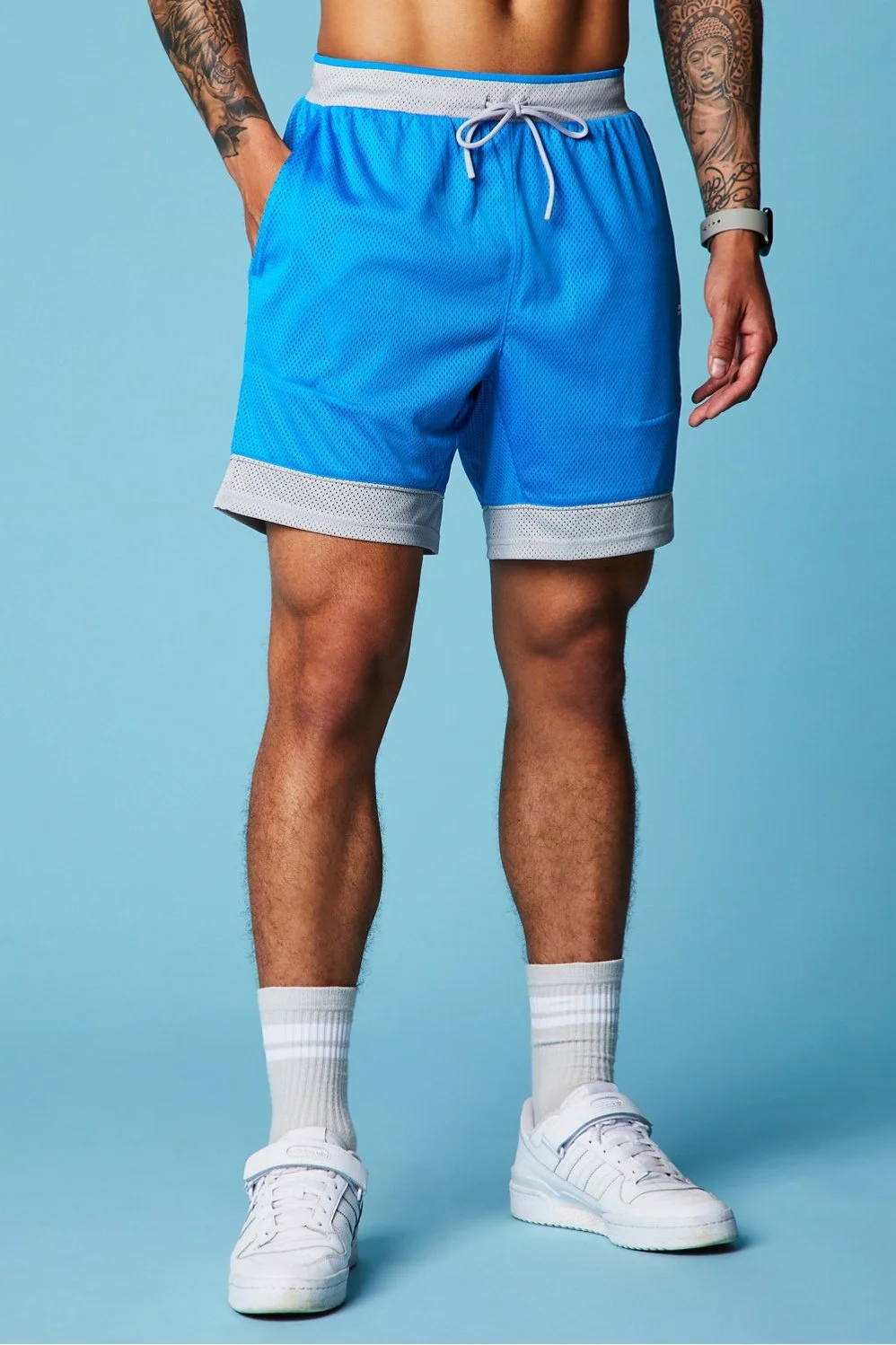 A man wearing blue athletic shorts with white trim, white socks, and white sneakers, standing against a light blue background.